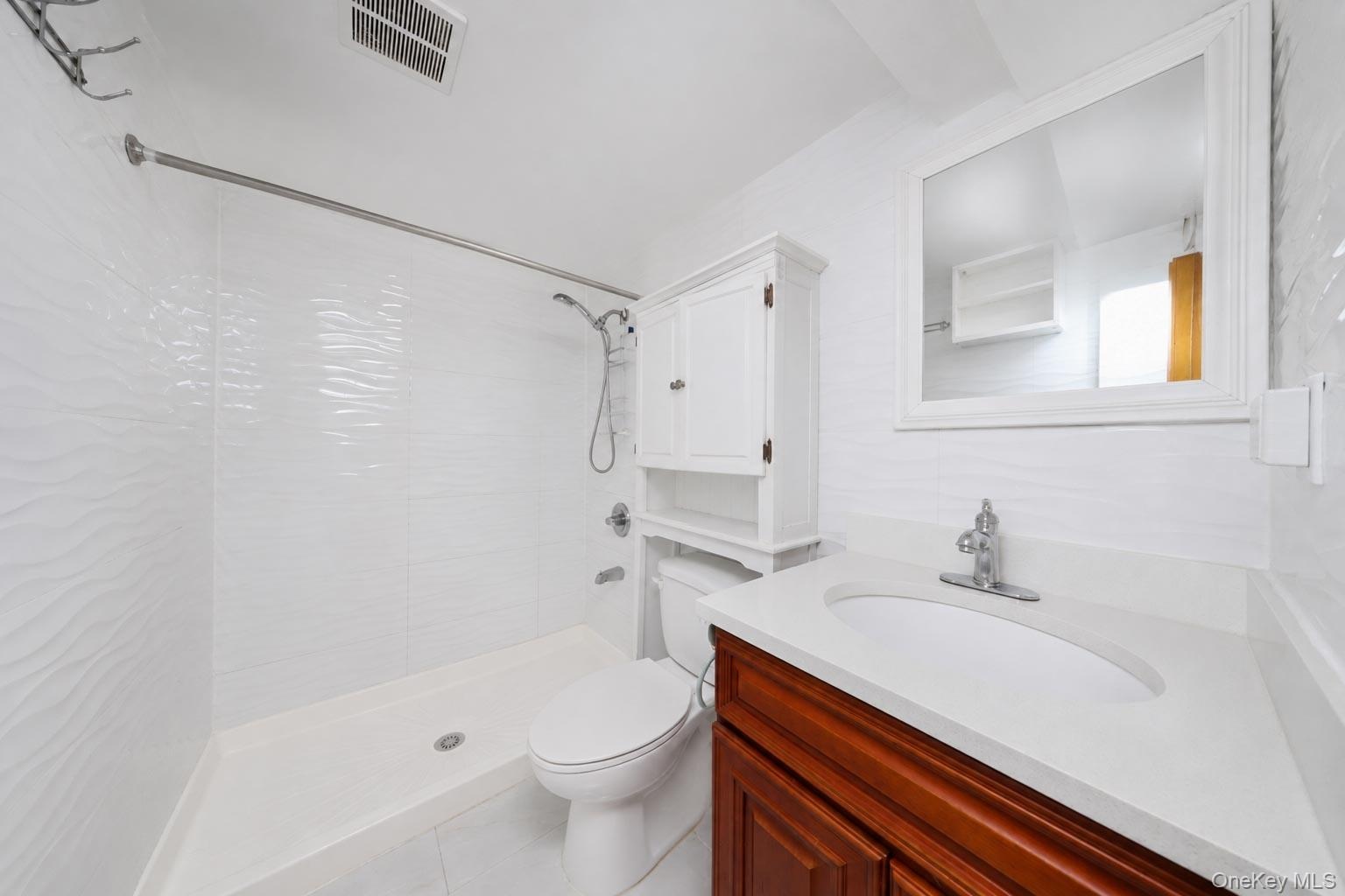 White textured shower tile, integrated shower system, white countertop with integrated sink, wood-finish vanity, and wall-mounted storage cabinet