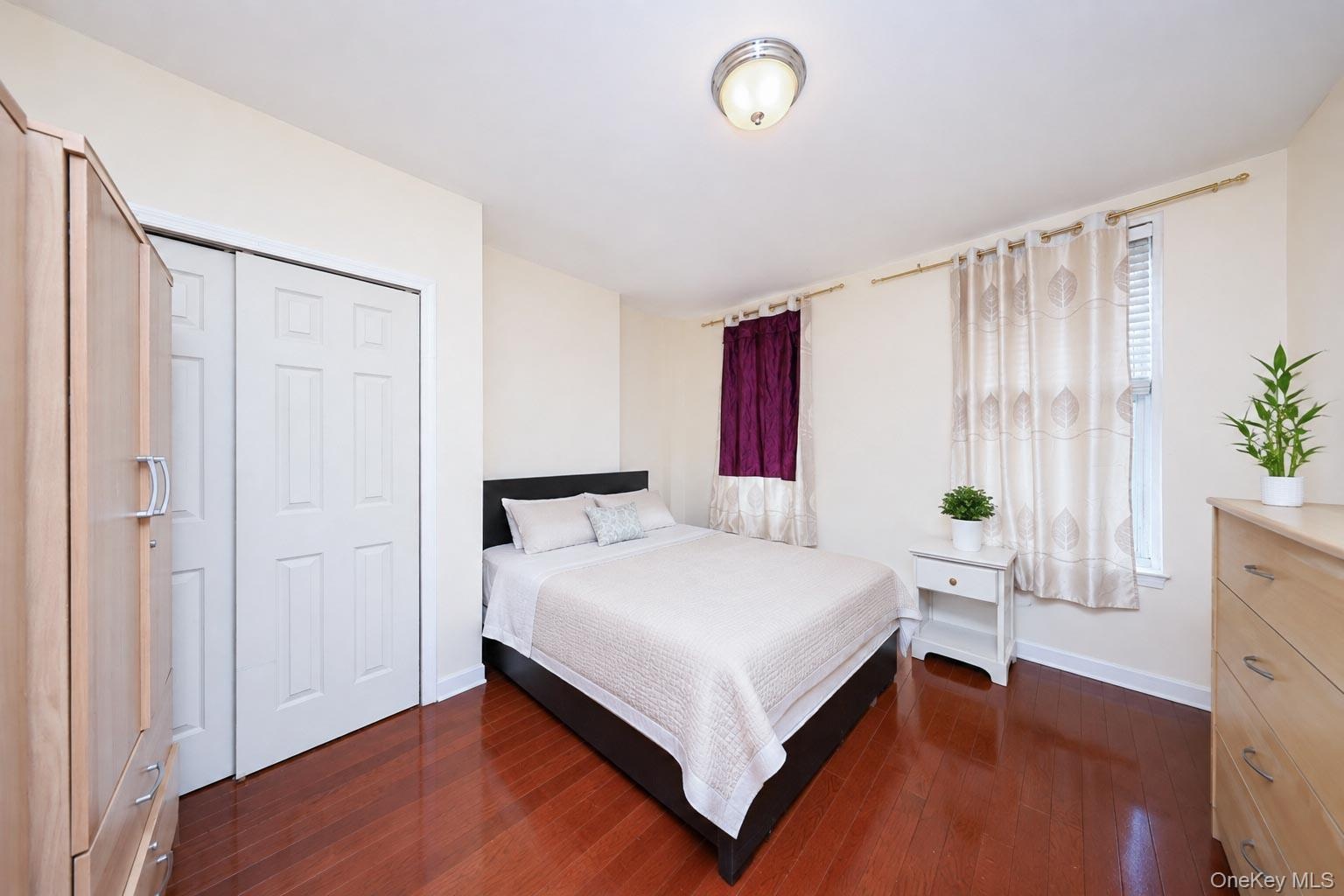 Spacious bedroom featuring rich wood-finish flooring, a queen-size bed with upholstered frame, dual windows with sheer and thermal curtains, white six