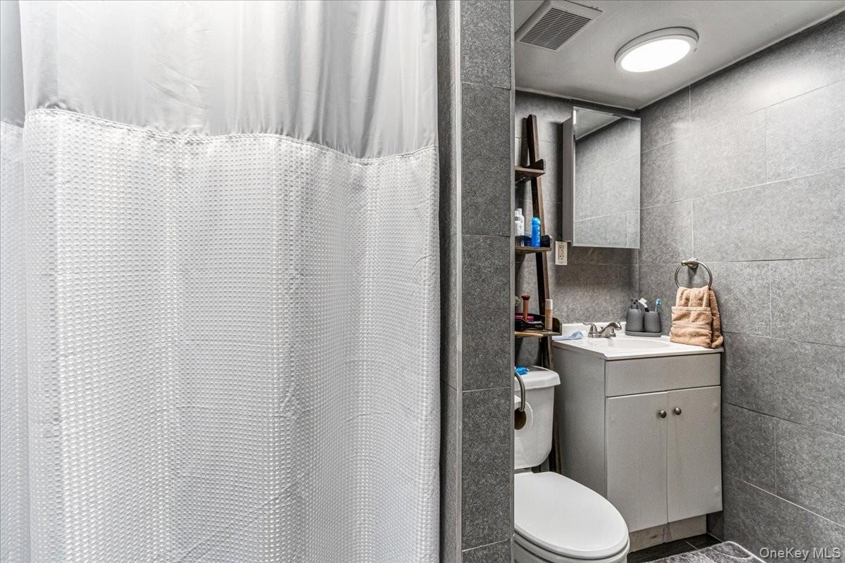 The bathroom is fully finished with modern grey wall tiling, a clean white vanity, and bright overhead lighting. This additional full bath includes a 