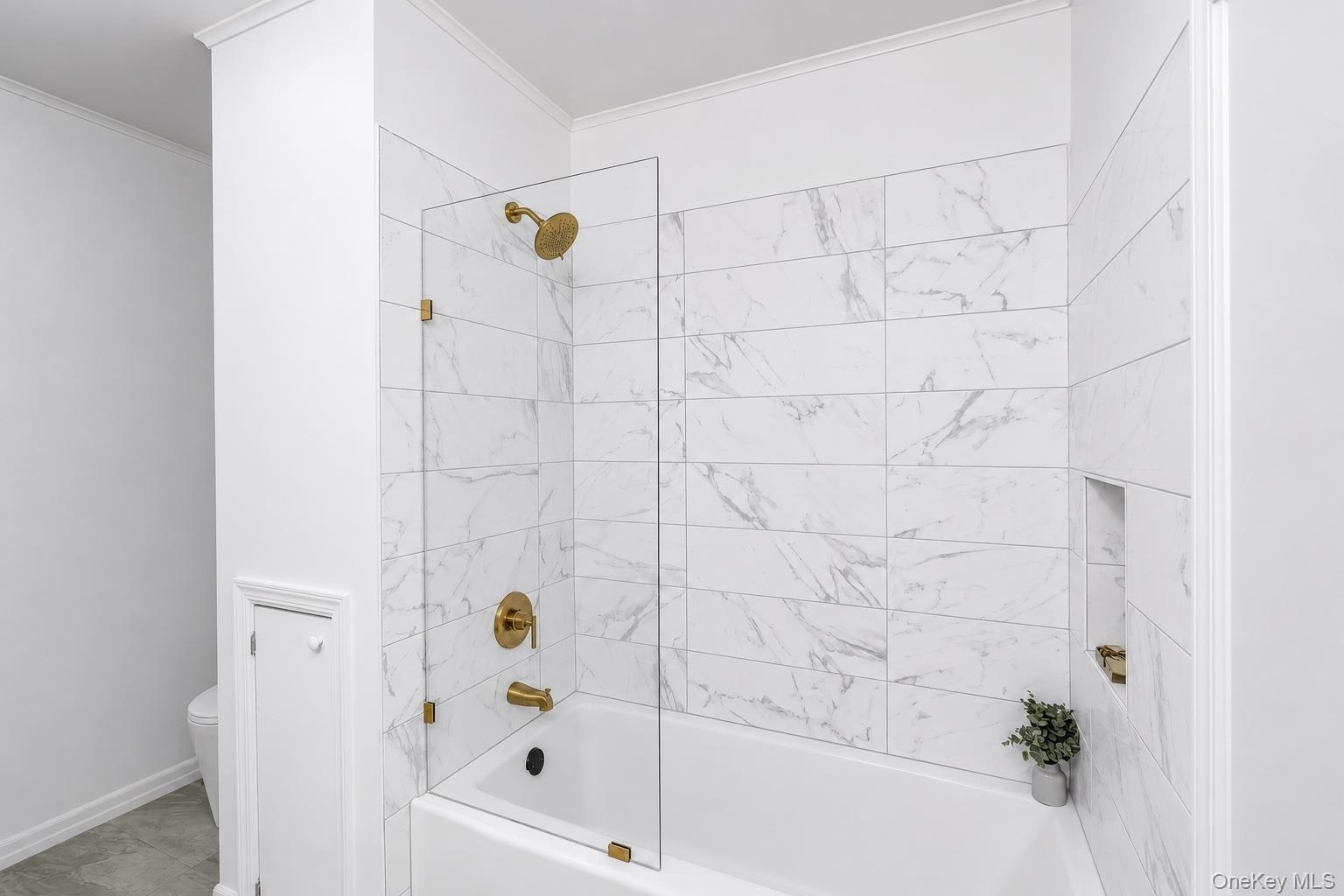 Bathtub with glass shower partition, gold-finish fixtures, and marble-look subway tile surround