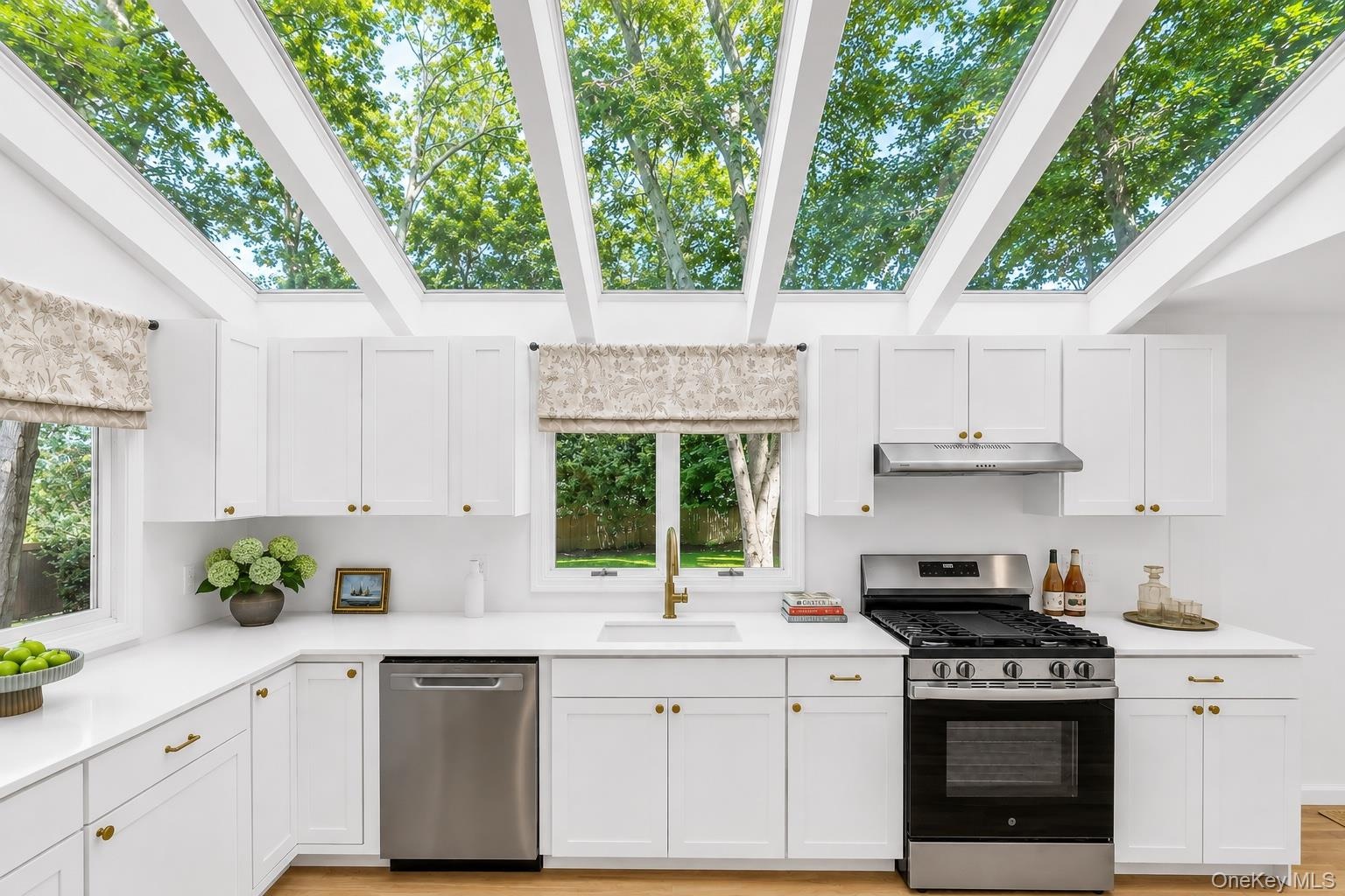 White cabinetry with brass hardware, stainless steel appliances, light wood-finish flooring, and a vaulted ceiling with exposed beams and skylights