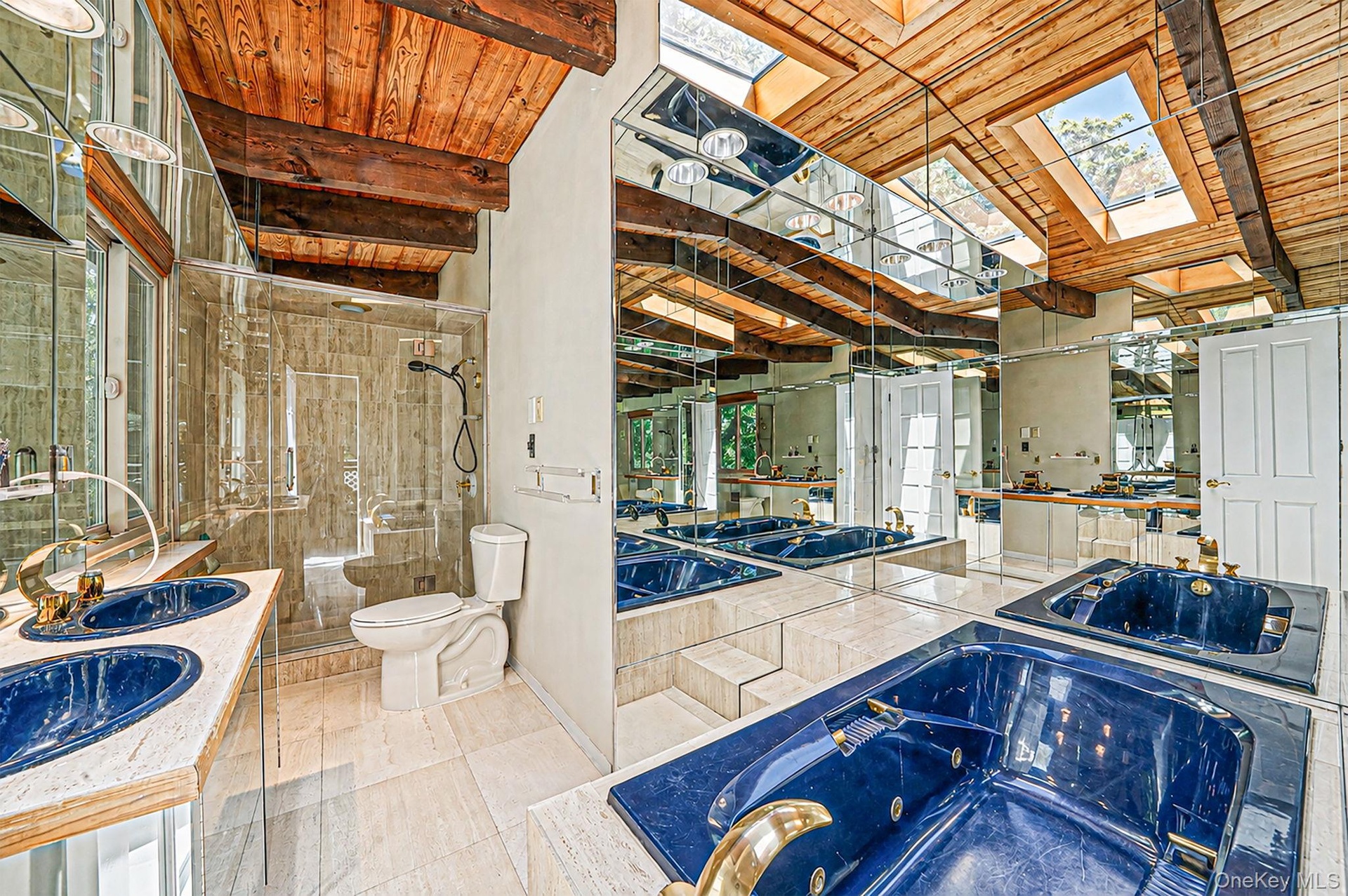 Wood-beamed vaulted ceiling with skylights, mirrored walls, dual integrated deep blue sinks, a walk-in shower with glass enclosure, and a built-in jet