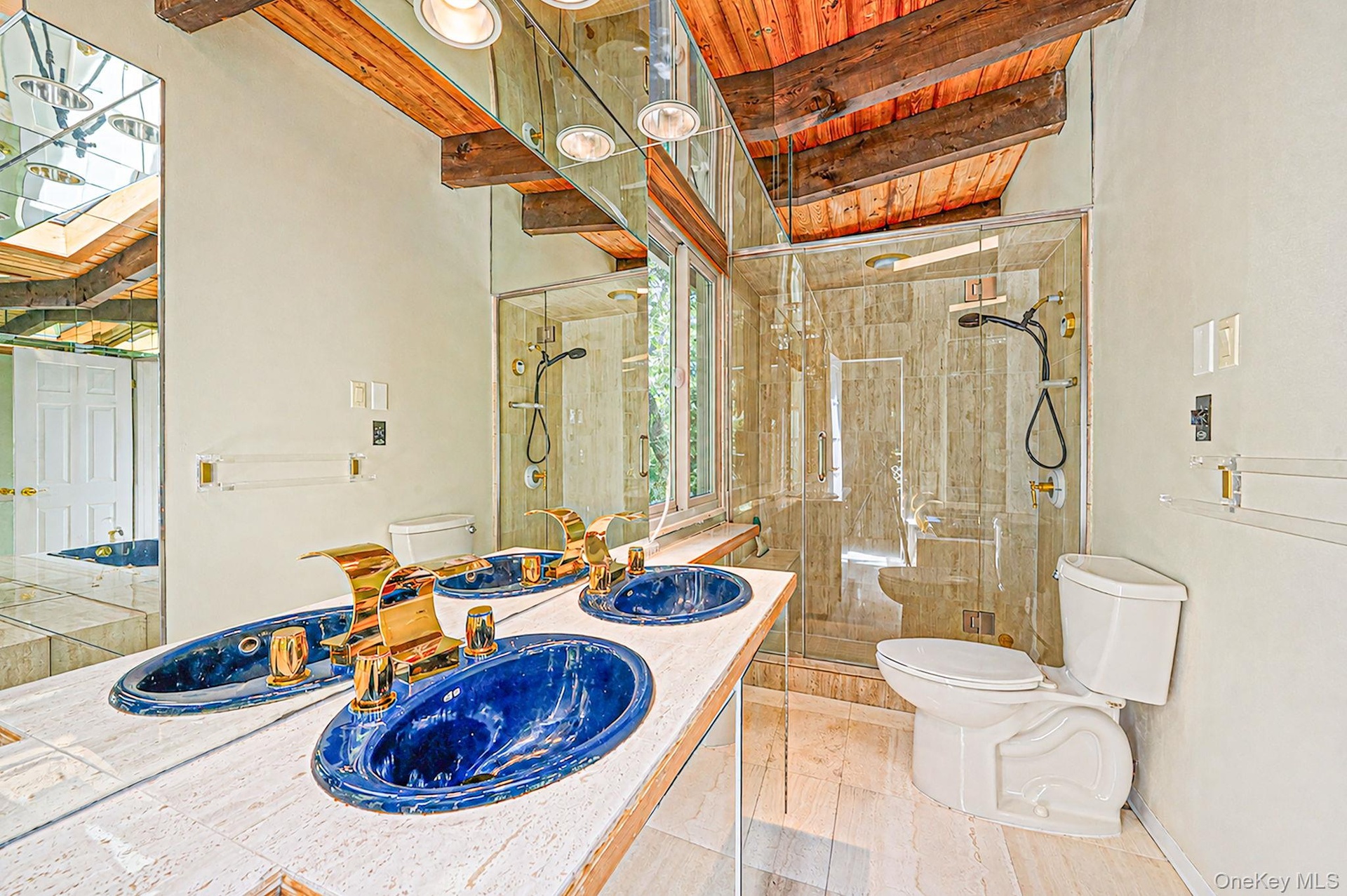 Dual vanity with blue vessel sinks and gold-tone fixtures, glass-enclosed shower, exposed wood beam ceiling, and skylight