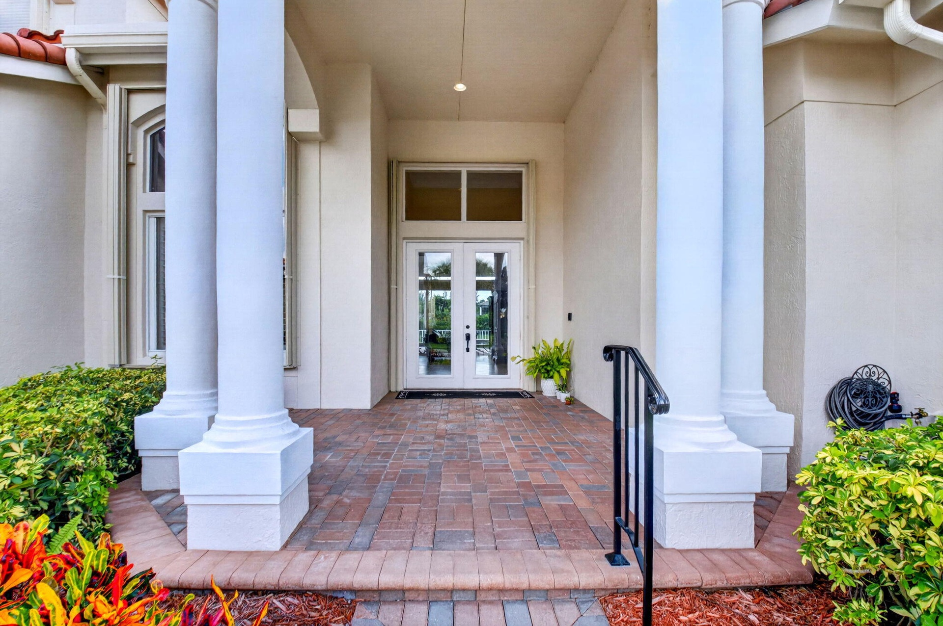 Portico entry featuring large columns, double glass-paneled doors, brick paver flooring, and a wrought iron handrail