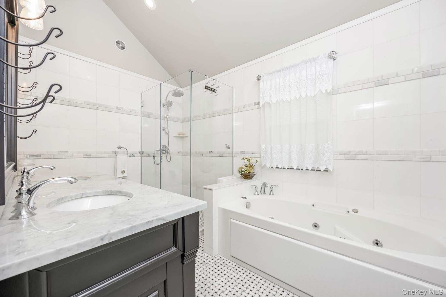 White tiled bathroom featuring a single vanity with stone countertop, glass-enclosed shower, built-in soaking tub, and black and white mosaic flooring