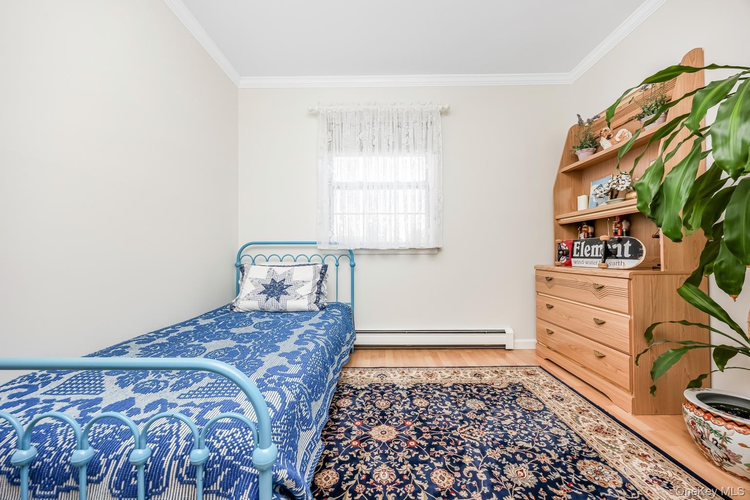 Bright bedroom featuring a metal bed frame, wood-finish dresser with integrated shelving and potted greenery, dark patterned area rug, and a single-hu