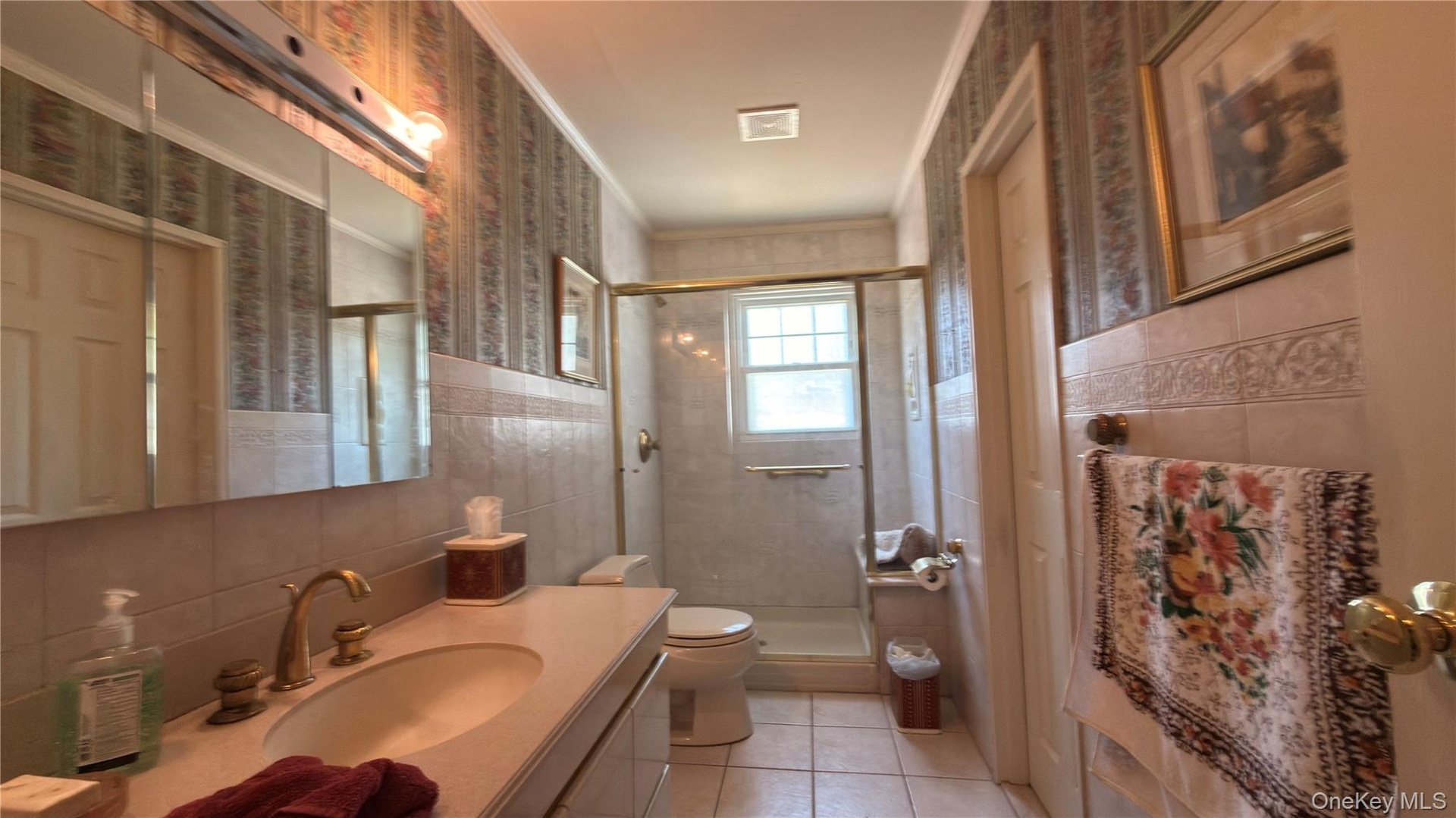 Vanity with integrated sink, brass fixtures, mirrored medicine cabinet, tiled shower with glass enclosure, and a window