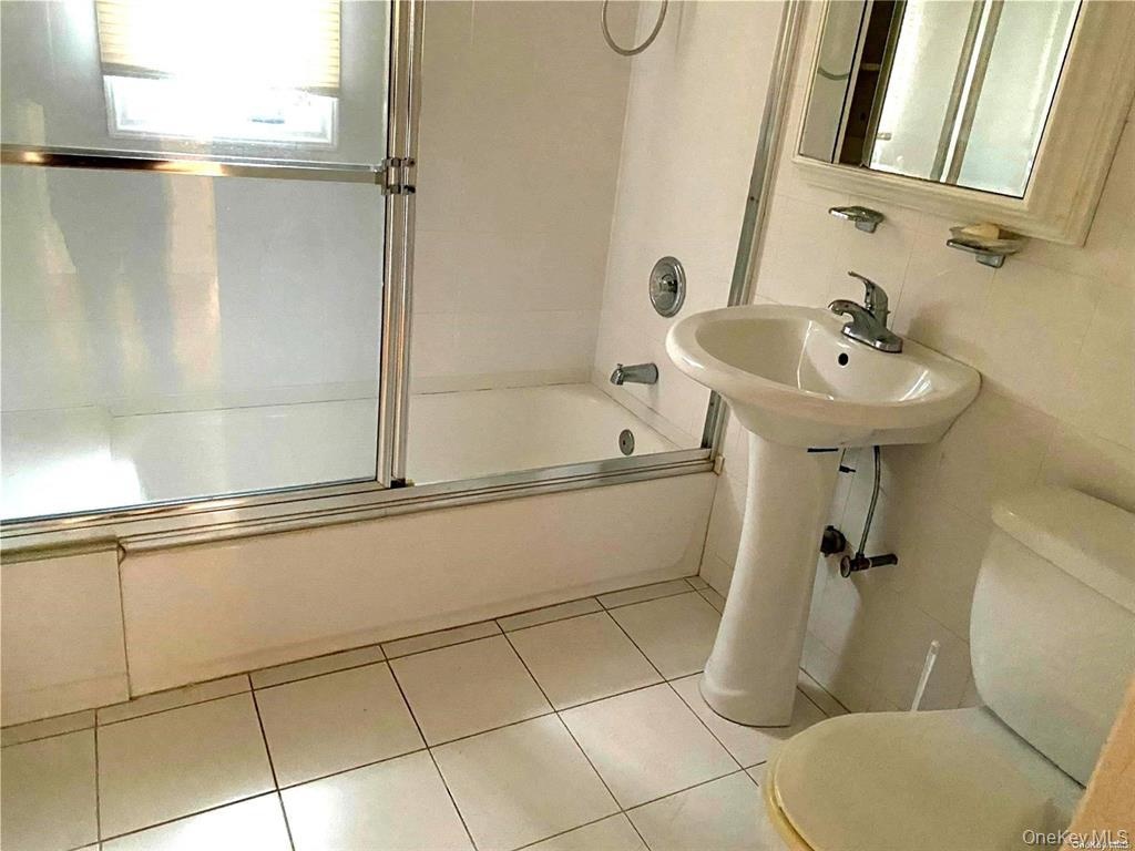 Pedestal sink with chrome faucet, white tiled flooring, bathtub with sliding glass enclosure, white wall tiling, and mirrored vanity cabinet