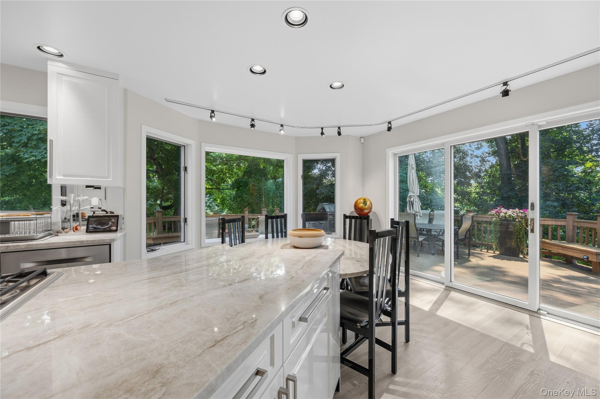 View from inside the gleeaming white kitchen with walls of windows and sliding glass doors to the wrap-around deck with another outside kitchen area.