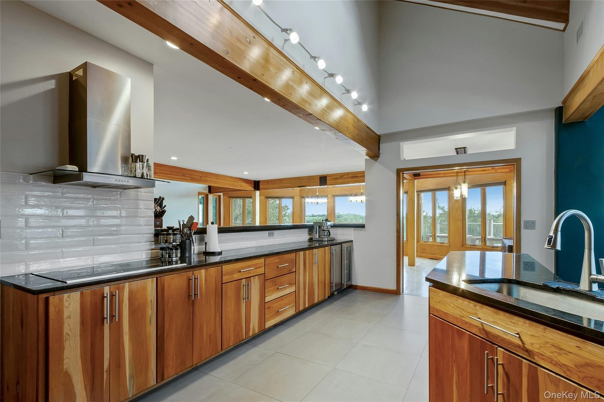 Vaulted ceiling with exposed wood beams, stainless steel range hood, natural wood cabinetry, black countertops, and a white subway tile backsplash