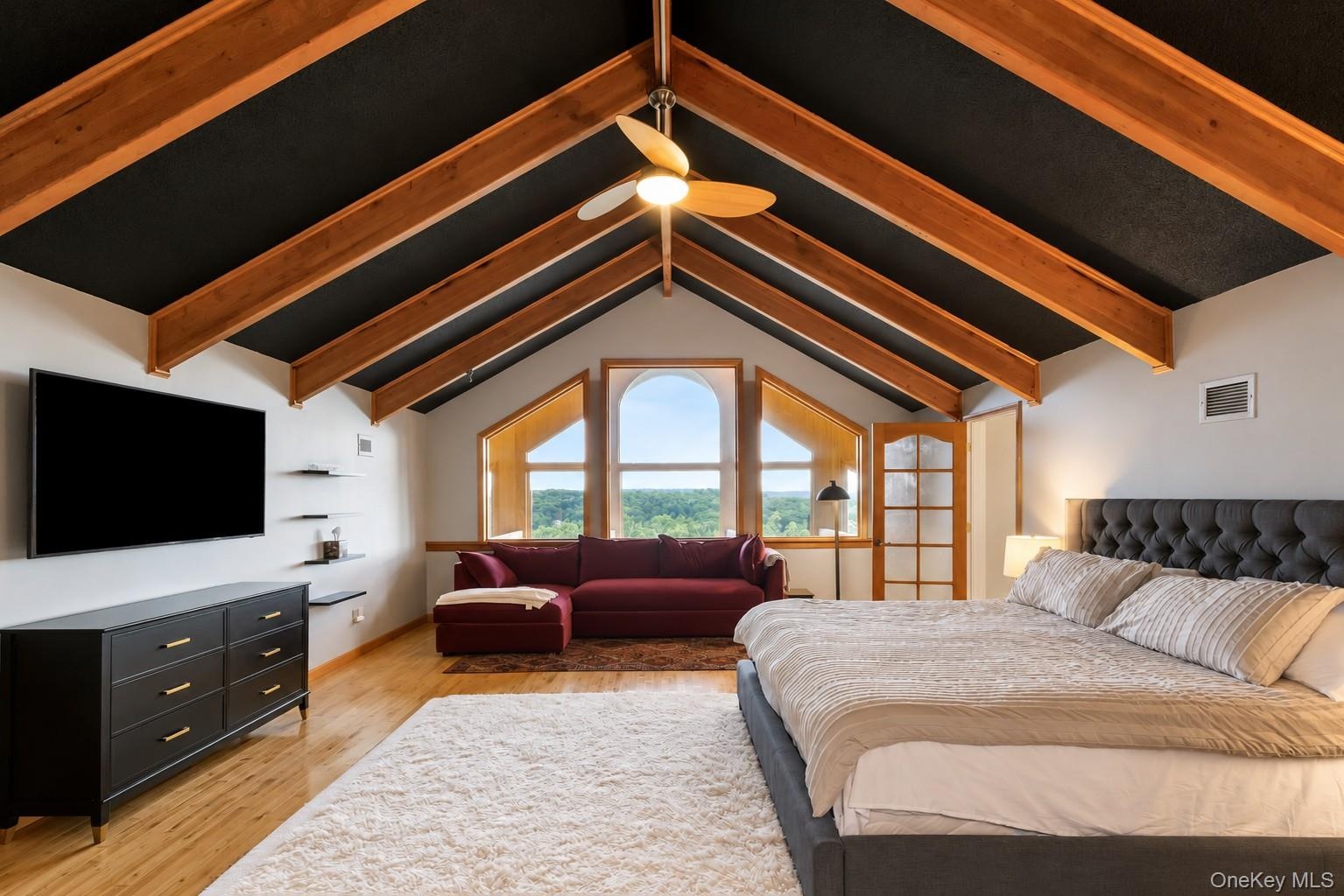 Vaulted ceiling with exposed wood beams, arched windows, light wood-finish flooring, and a ceiling fan