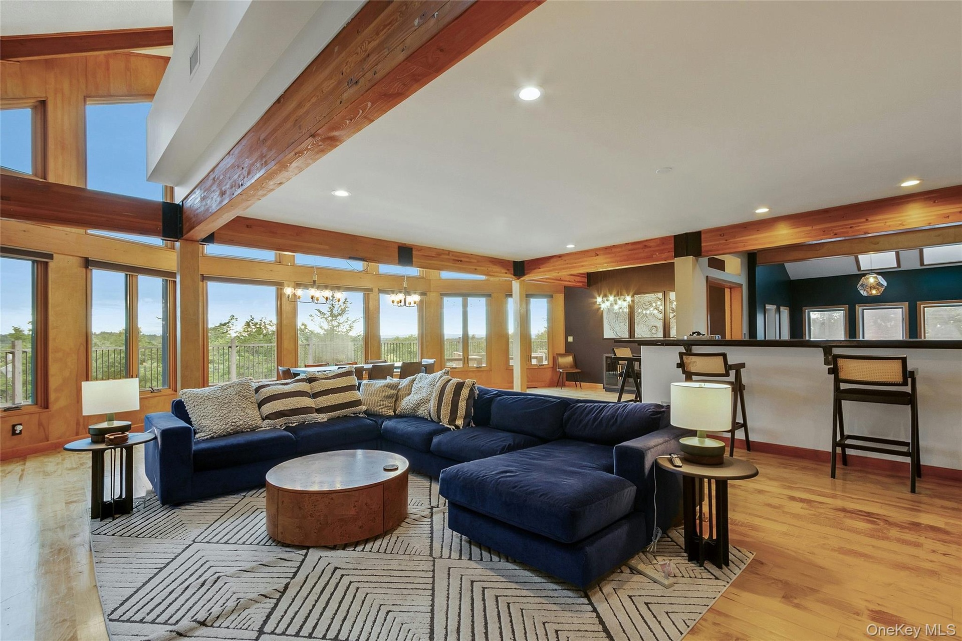 Expansive living space featuring exposed wood beams, wood-finish flooring, a full-height window wall, recessed lighting, and a built-in bar with count
