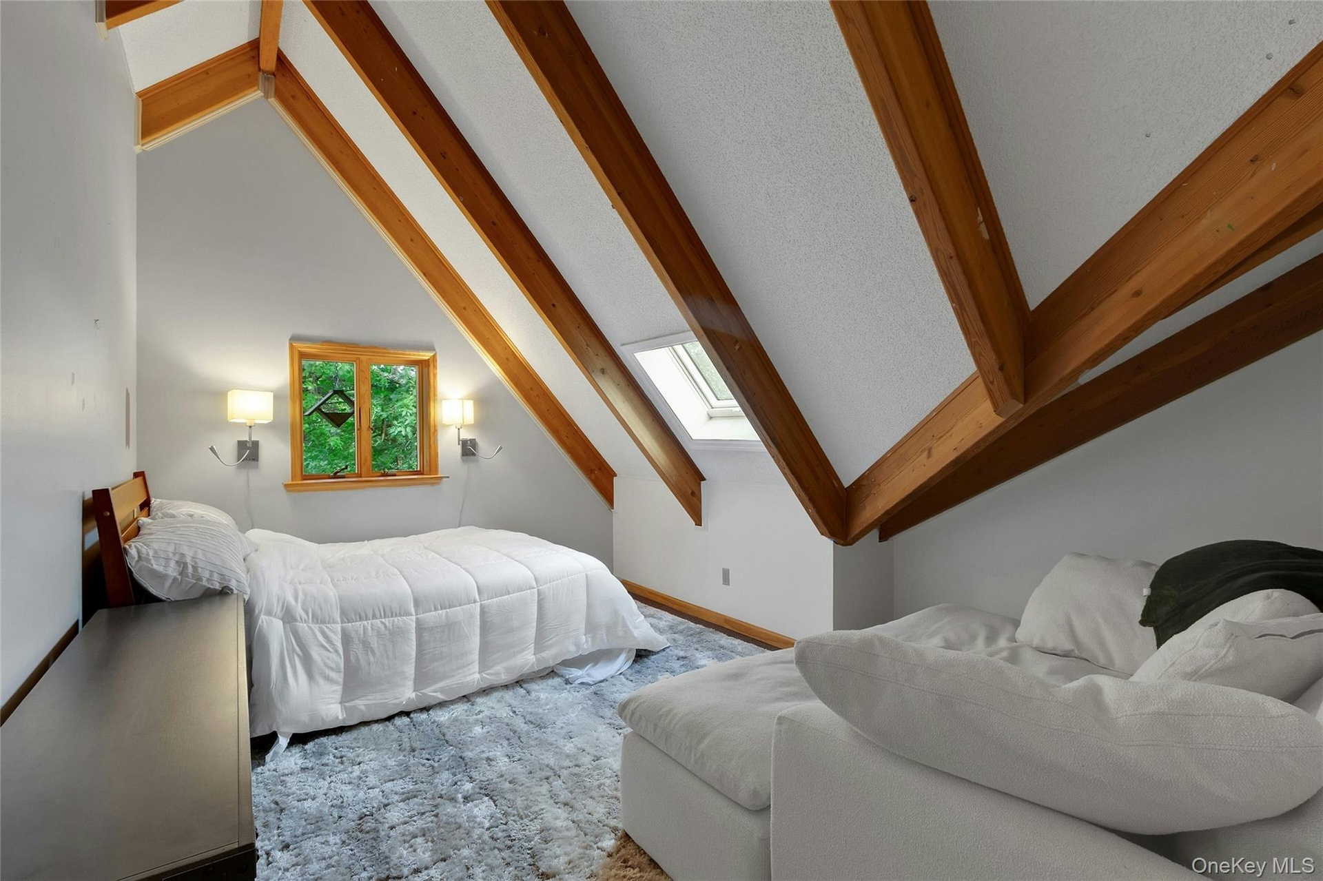 Vaulted ceiling with exposed wood beams, wood-frame window, two wall-mounted sconces, and a skylight