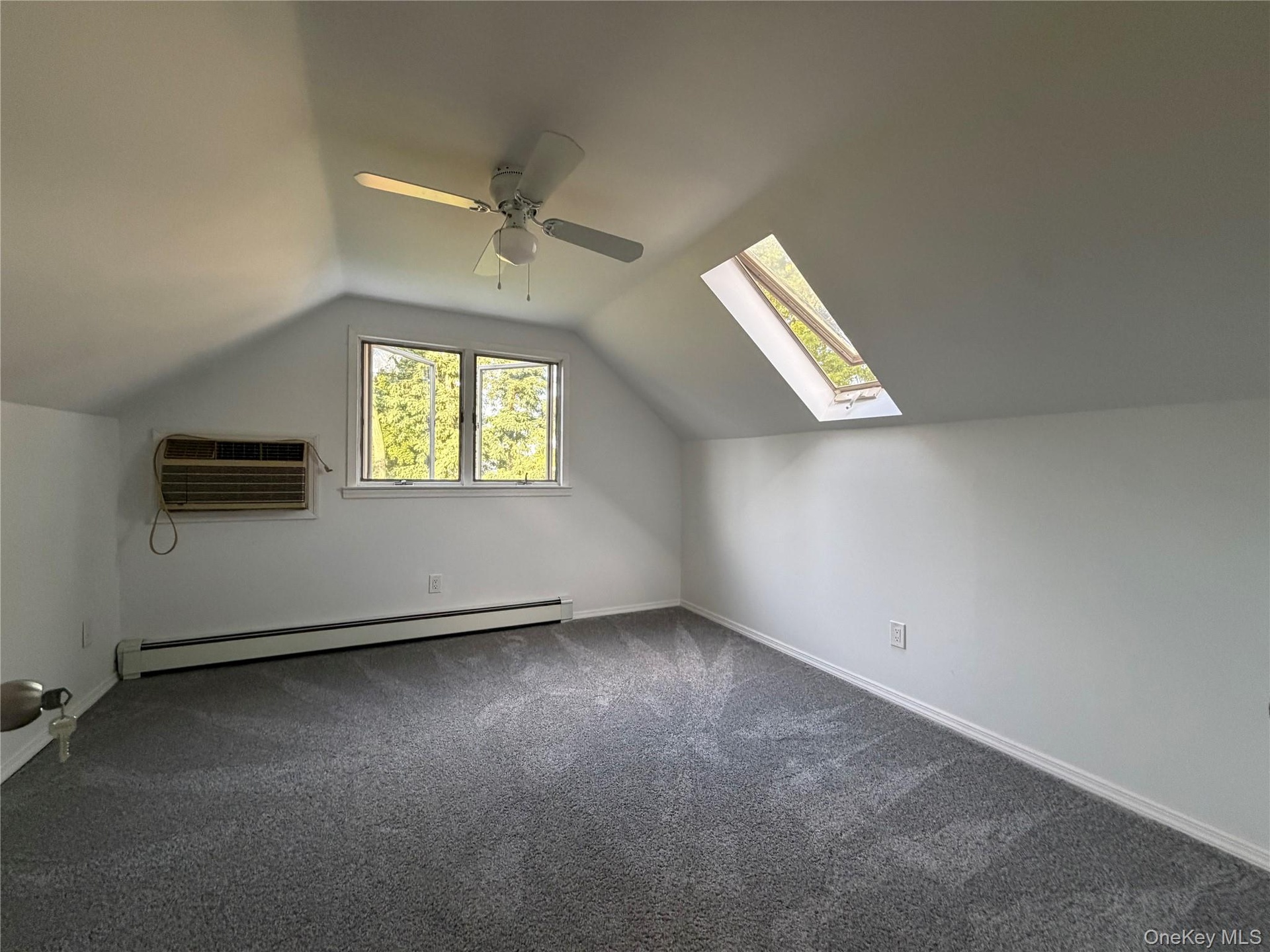 Vaulted ceiling with a ceiling fan, skylight, wall-mounted air conditioning unit, baseboard heating, and gray carpeting