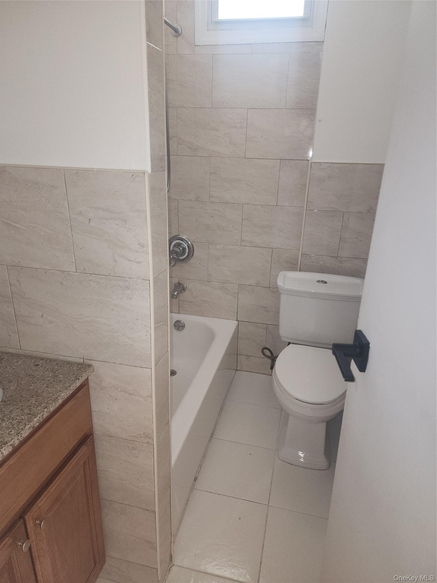 Full bathroom featuring a white tub, light-toned wall tiling, white toilet, and a vanity with a light-colored countertop