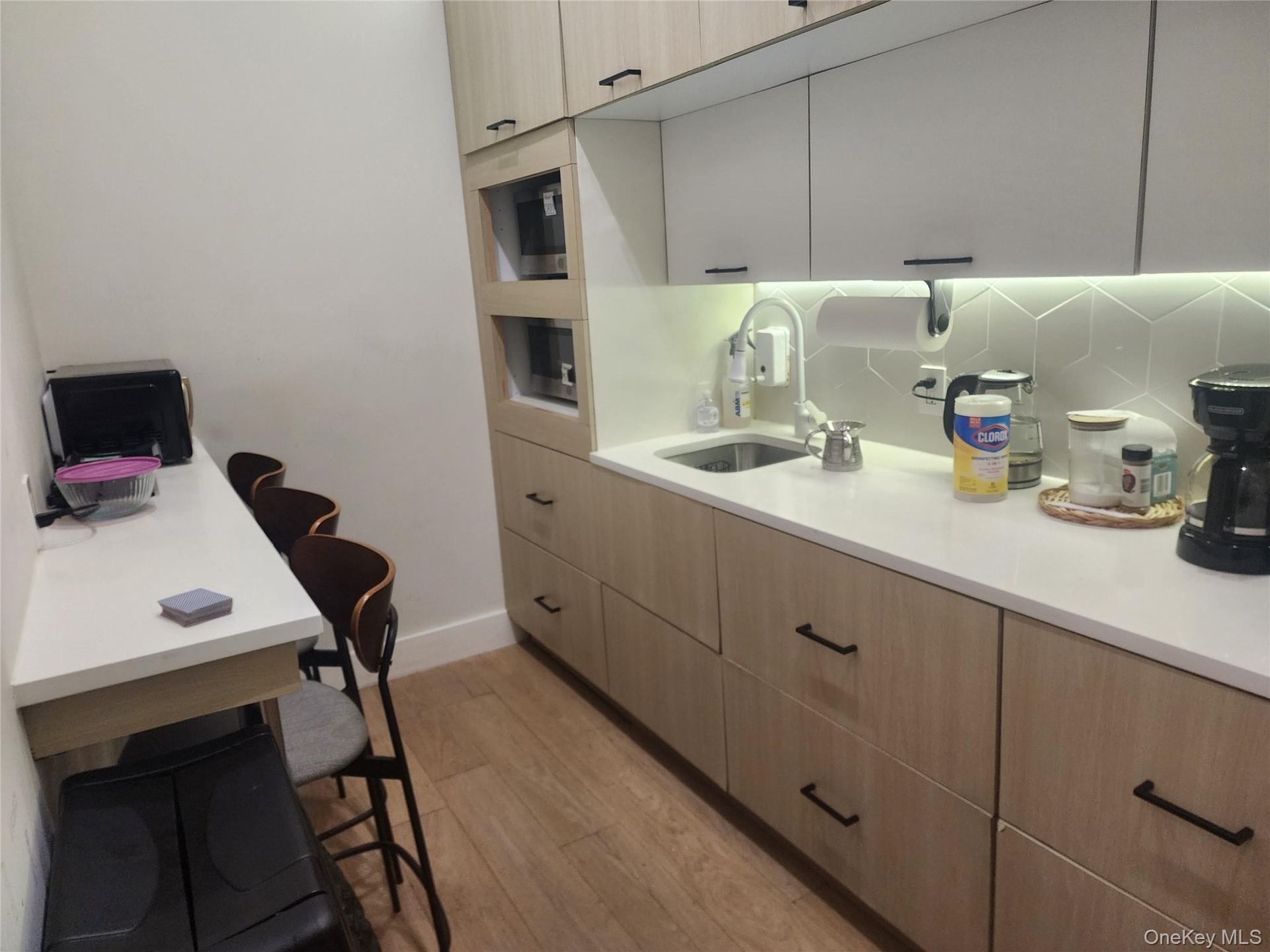 Compact kitchen area featuring wood-finish flooring, light-toned cabinetry with dark hardware, built-in microwave, white solid surface countertops, an