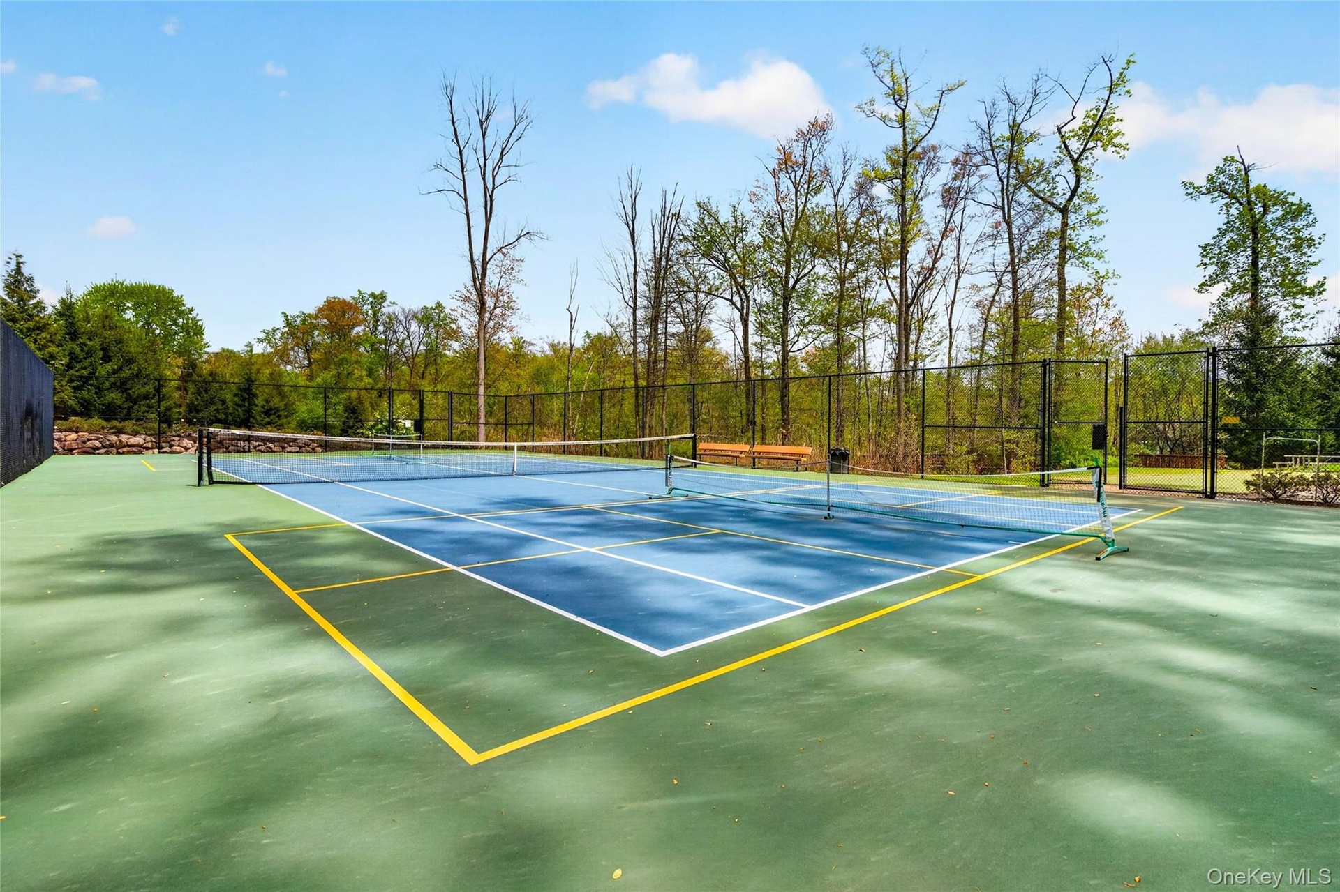 Hard-surface tennis court with a blue playing area and green perimeter, surrounded by black fencing