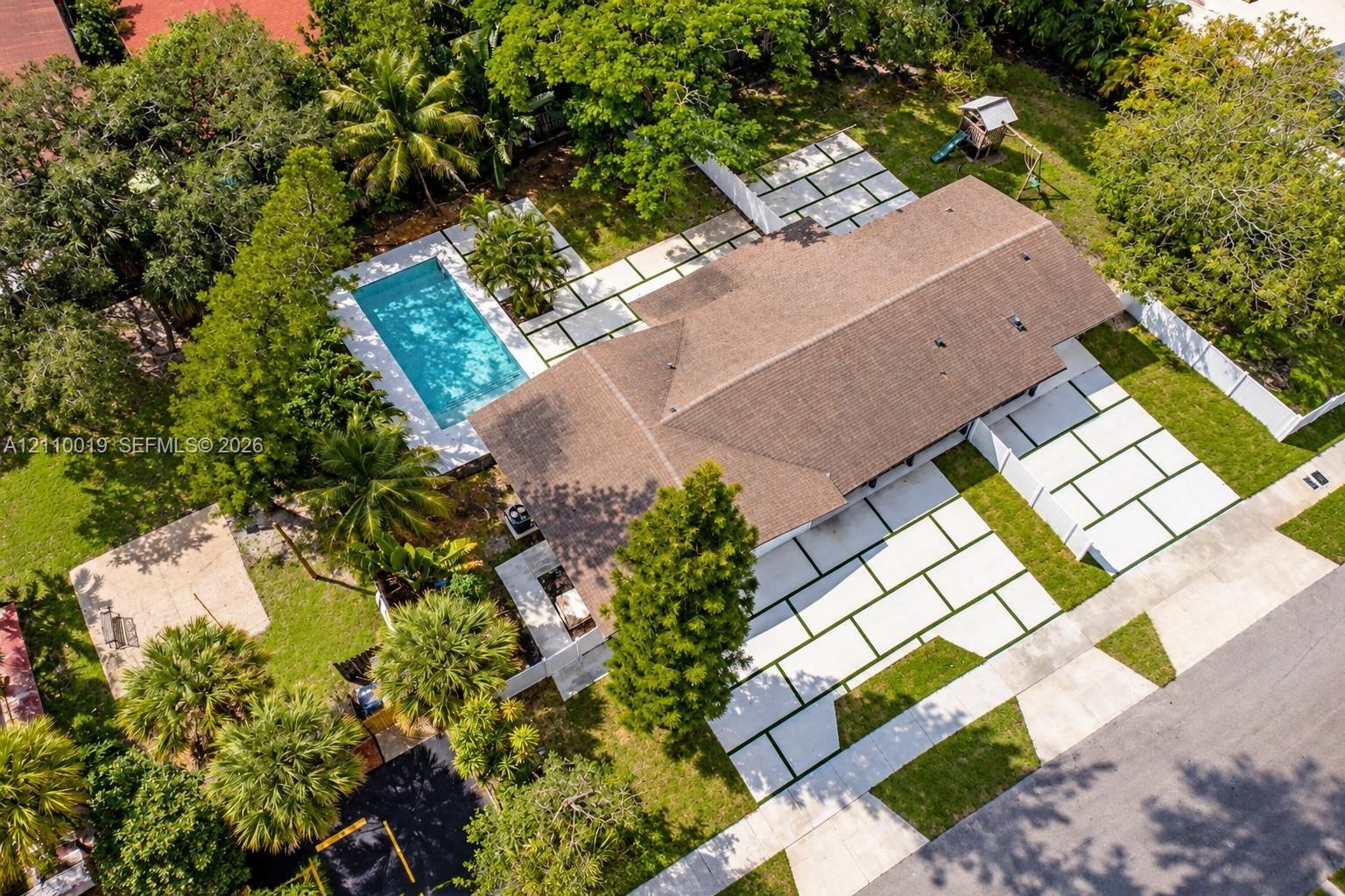 Aerial perspective of a property featuring an in-ground swimming pool, extensive paver hardscaping with geometric inlay, mature landscape, and a fence