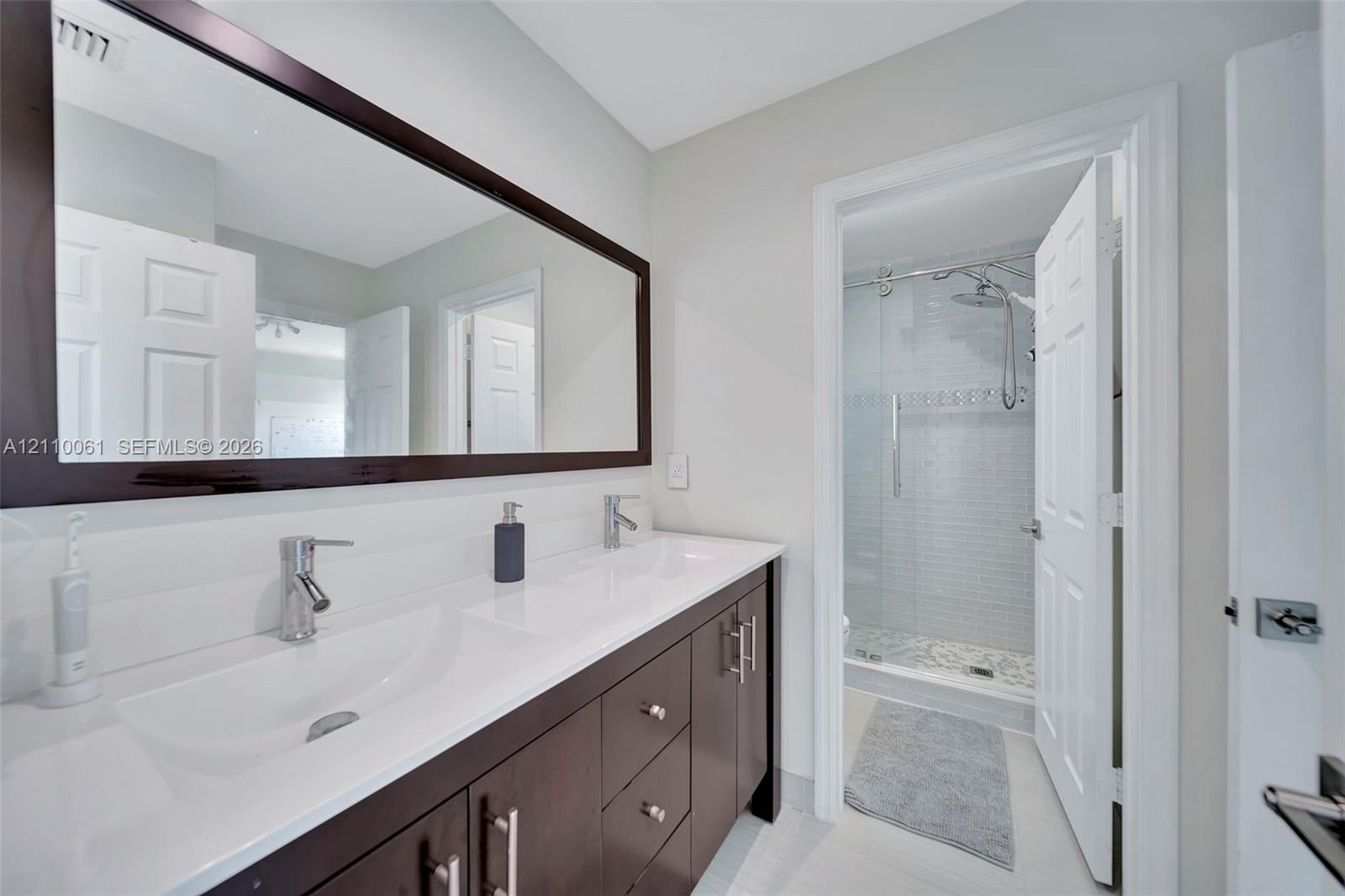 Dual-sink vanity with dark wood-finish cabinetry, solid-surface countertop, full-width mirror, and walk-in glass shower enclosure