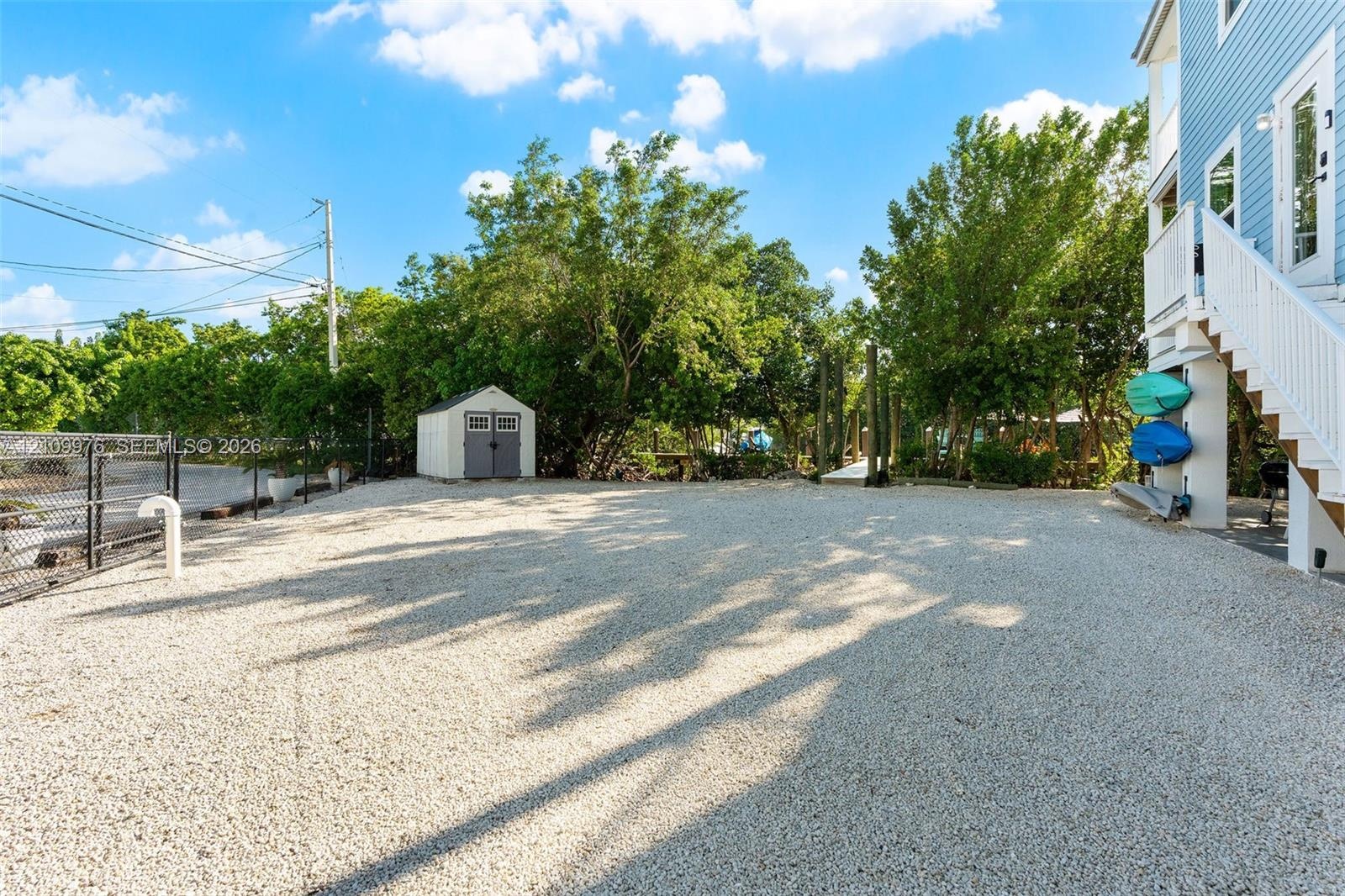 Gravel-surfaced exterior lot featuring a detached storage shed, perimeter chain-link fencing, and elevated home foundation