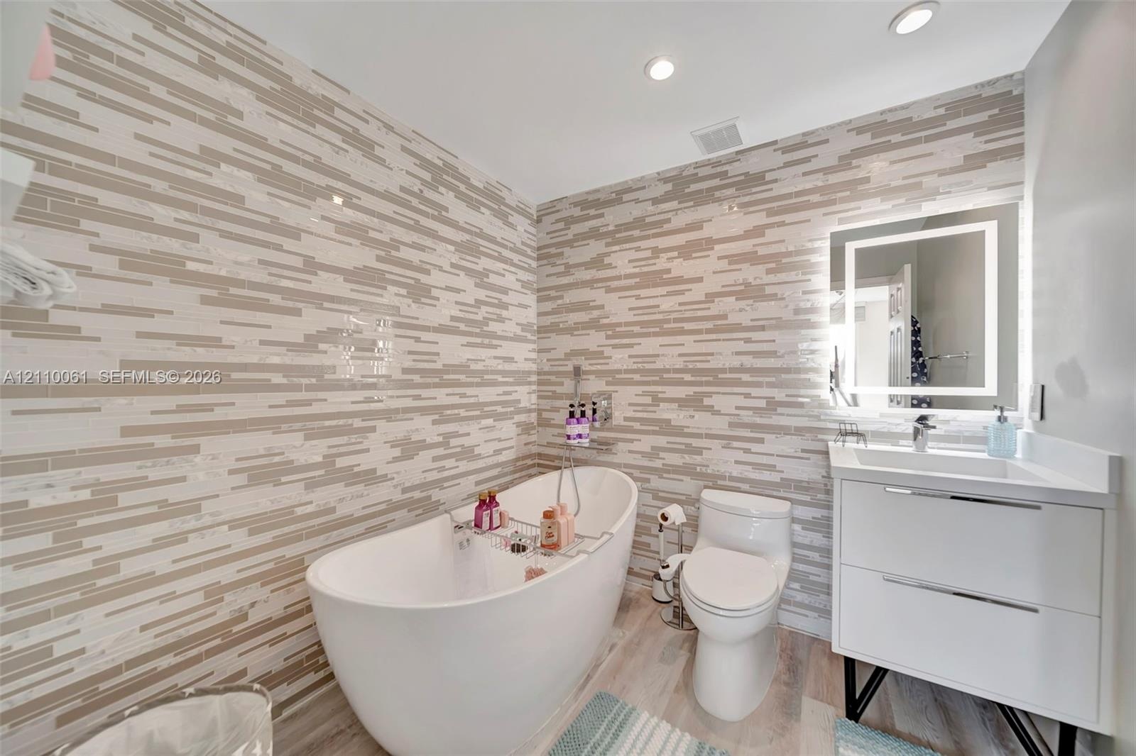 Bathroom featuring floor-to-ceiling multi-tonal linear tile walls, a freestanding soaking tub with handheld sprayer, a vanity with a white countertop 