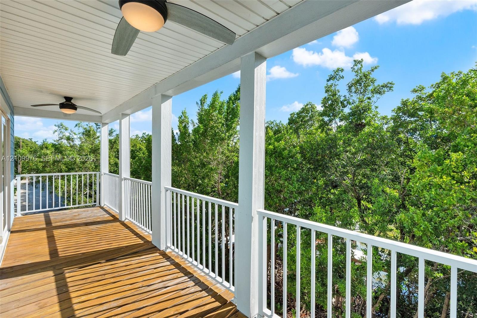 Covered balcony featuring wood decking, white baluster railings, dual ceiling fans with integrated lighting, and a white beadboard ceiling