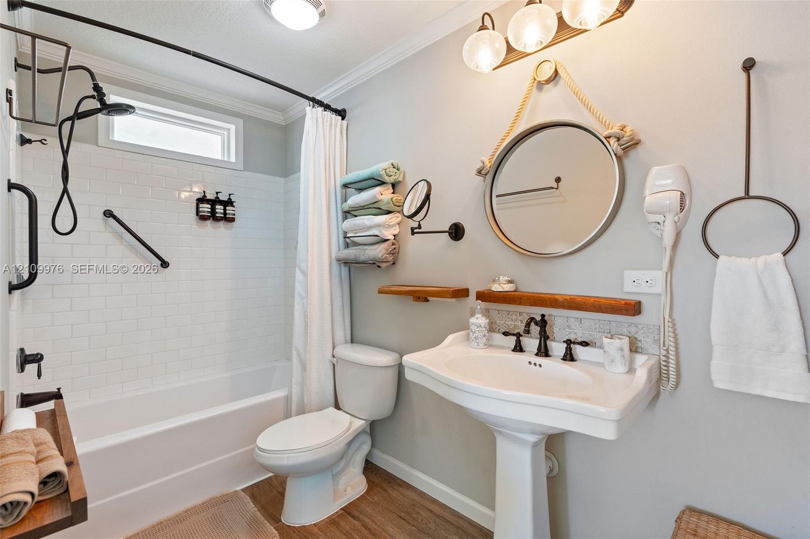 Bathroom featuring white subway tile tub surround, pedestal sink with multi-handle oil-rubbed bronze faucet, round wall-mounted mirror, wood-finish fl
