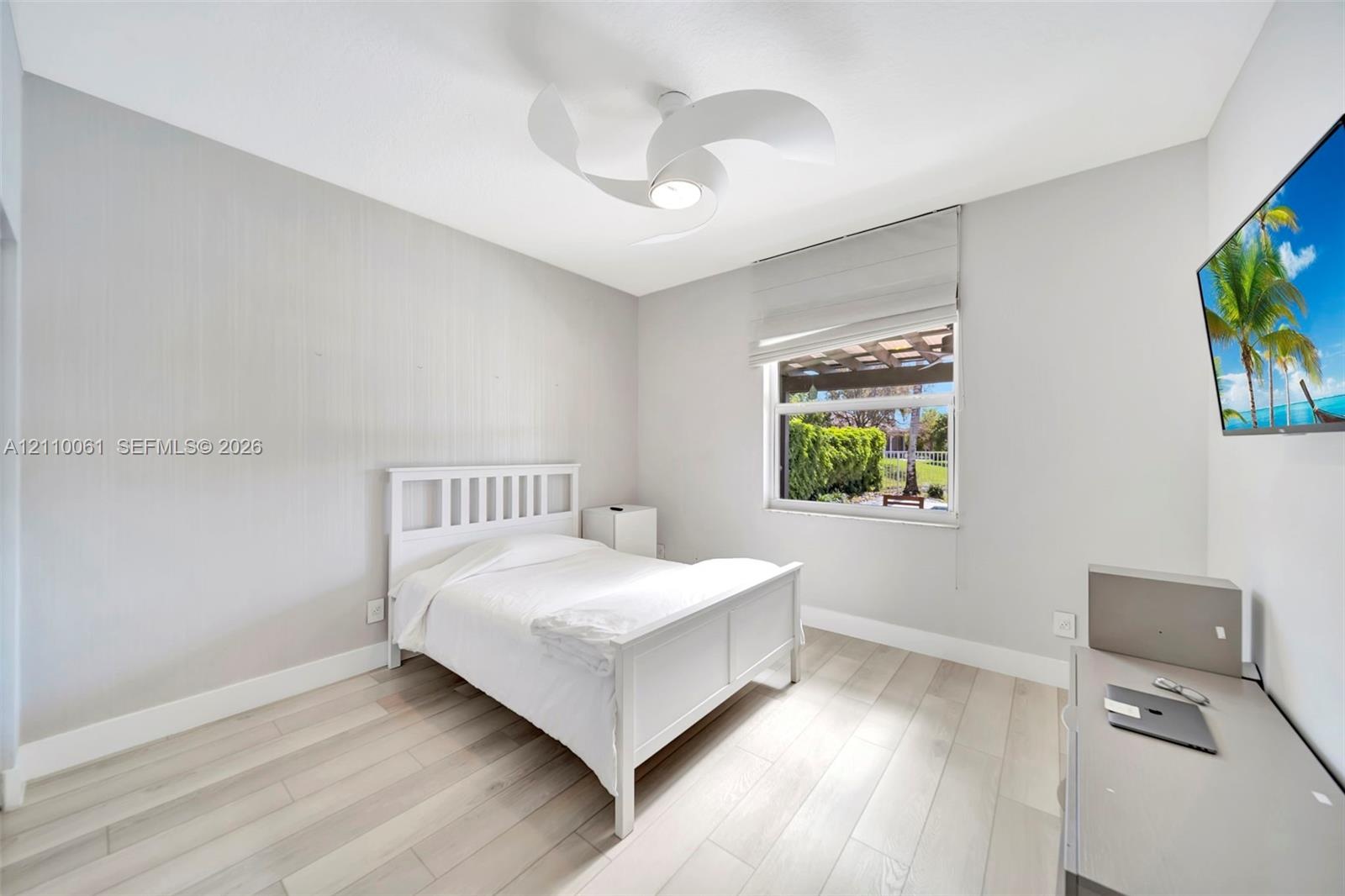 Wood-finish flooring, modern three-blade ceiling fan with integrated lighting, and neutral wall tones complemented by a window featuring a Roman shade
