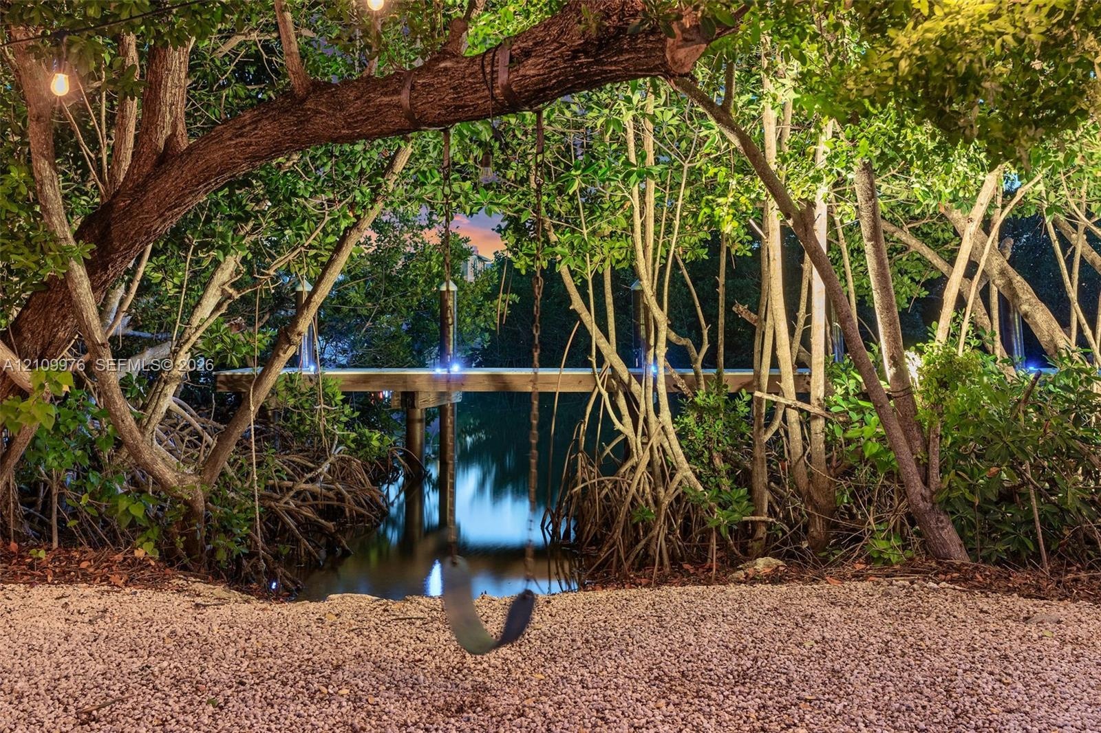 Waterfront grounds featuring a private wood dock, pebble-surfaced shoreline, mature coastal trees, and exterior string lighting
