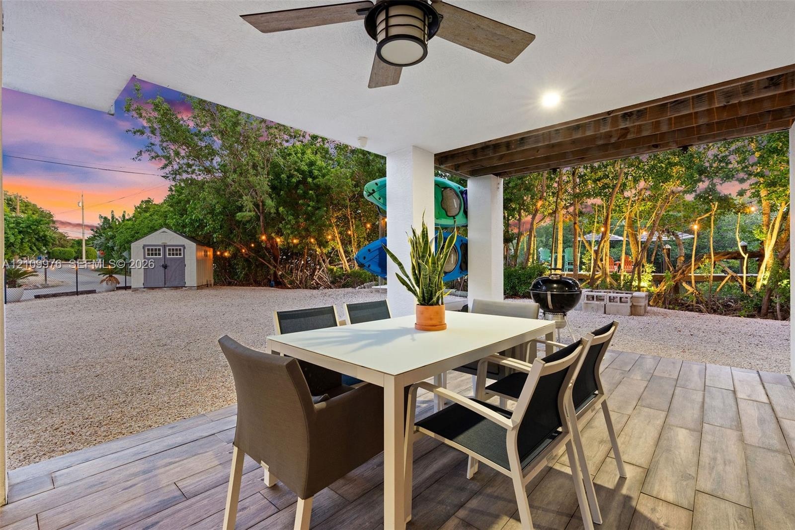 Covered patio featuring wood-finish tile flooring, ceiling fan, and exposed wood-beam detailing, opening to a gravel courtyard with a storage structur