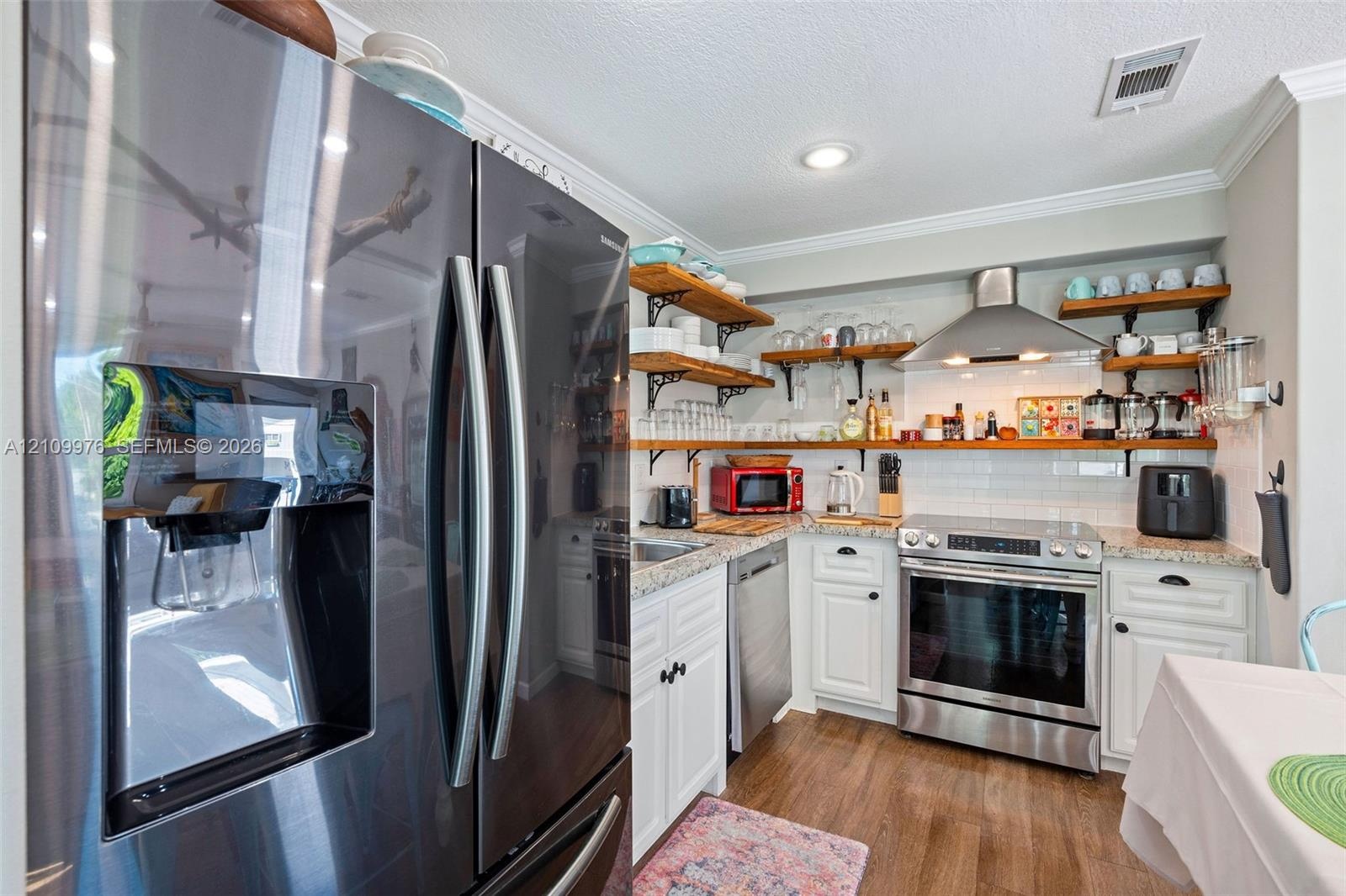 Kitchen featuring stainless steel appliances, stone-look countertops, white cabinetry with crown molding, natural wood-finish open shelving, and a whi