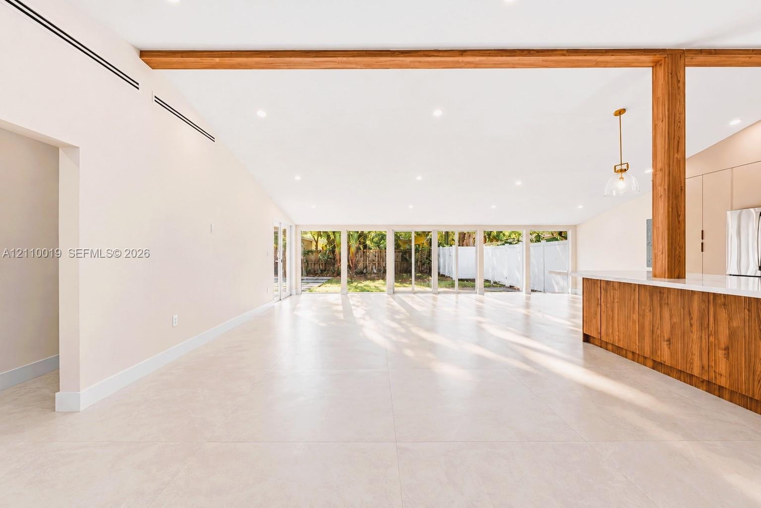 Open-concept interior featuring large-format tile flooring, exposed wood ceiling beams, floor-to-ceiling glass panel windows, and a wood-finish kitche