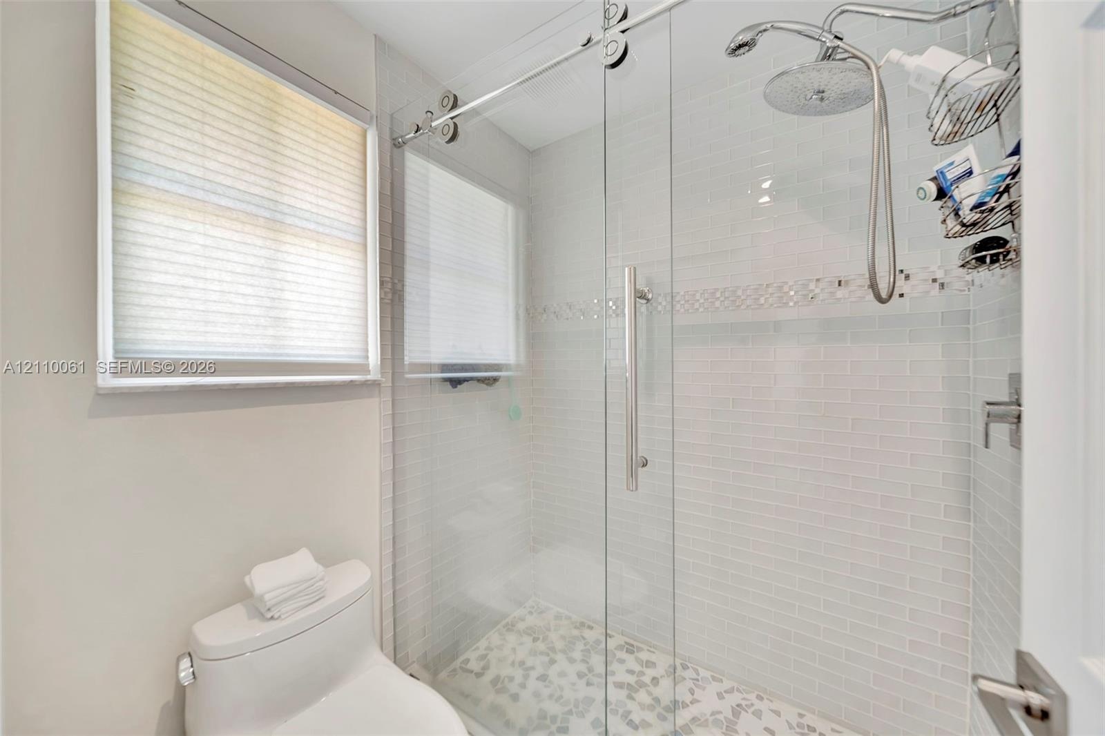 Walk-in shower featuring a glass sliding door, polished metal rain showerhead with handheld attachment, horizontal white tile walls with a decorative 