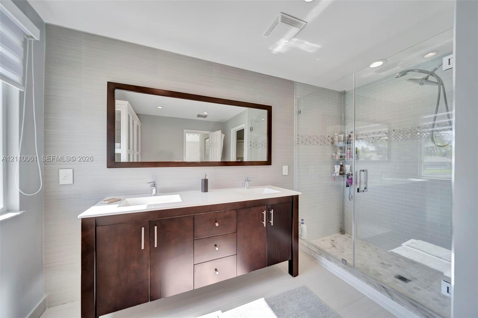 Double vanity with rectangular undermount sinks and light-toned solid surface countertop, wood-finish cabinetry with metallic hardware, and walk-in gl