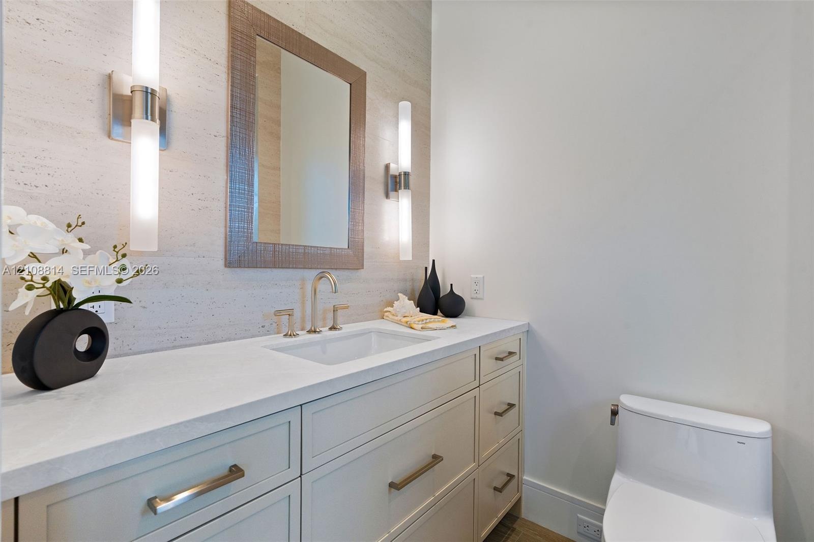 Custom vanity with quartz countertop, undermount sink, stone-finish wall cladding, dual vertical wall-mounted light fixtures, and framed mirror