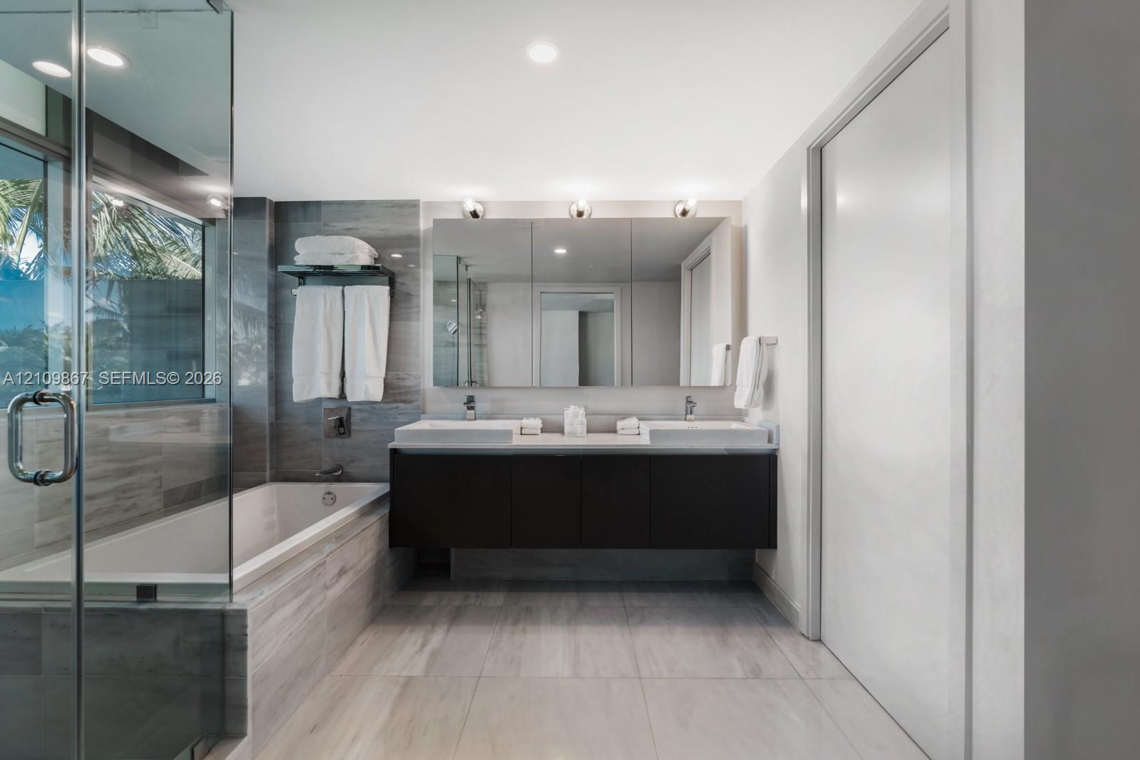 Floating dual-sink vanity with a countertop, expansive wall-mounted mirror, glass-enclosed soaking tub, and tile flooring