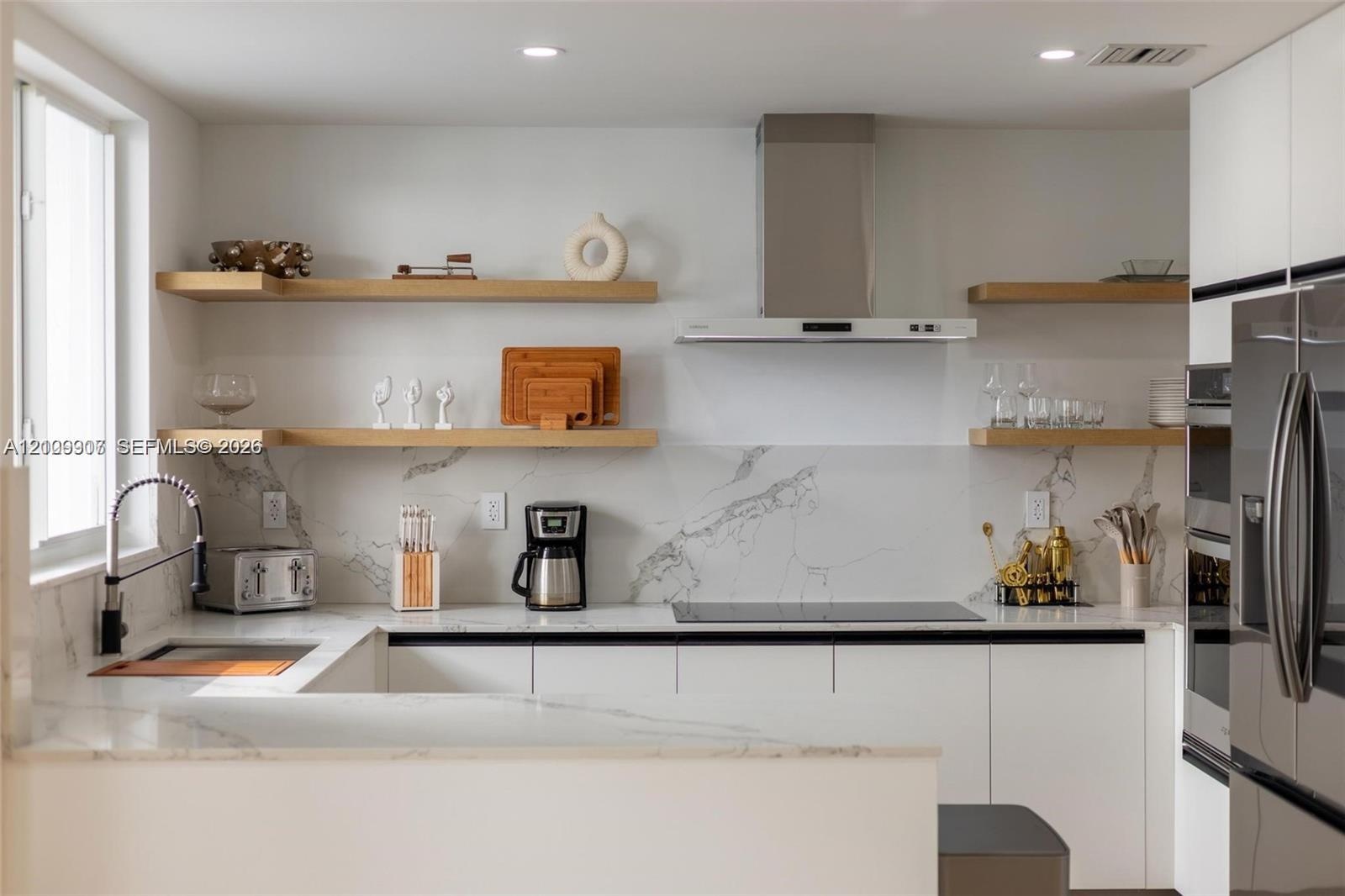 Modern kitchen featuring white flat-panel cabinetry, light-toned wood floating shelves, stone-surface countertops, full-height stone backsplash, and a