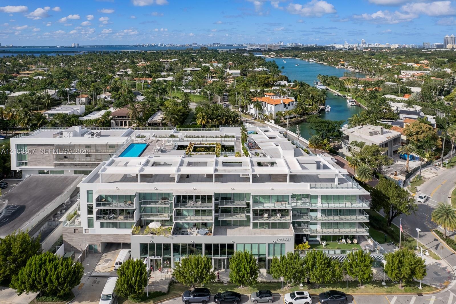 Multi-story residential complex featuring glass-railed balconies, a rooftop swimming pool, modern concrete exterior architecture, and mature landscapi
