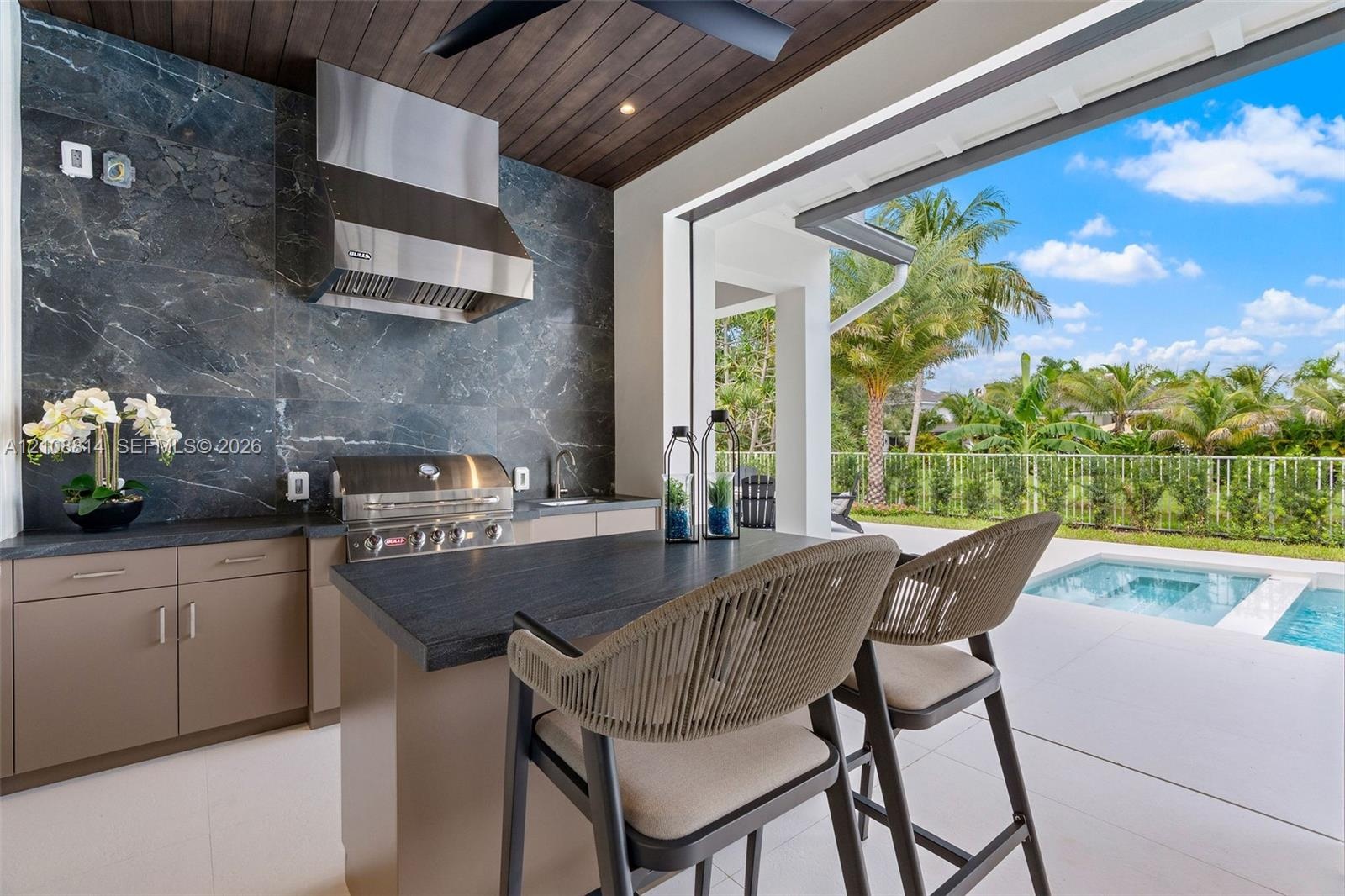 Covered outdoor kitchen featuring a dark stone backsplash, stainless steel range hood, integrated gas grill, flat-panel cabinetry, and dark-toned ston