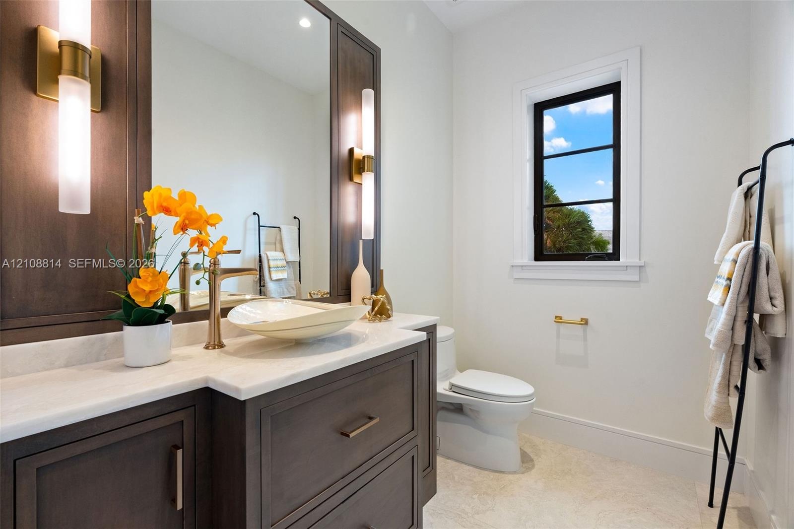 Dark wood vanity featuring a light-toned stone countertop, vessel sink, brass-finish hardware, integrated vertical sconces, and a multi-pane window