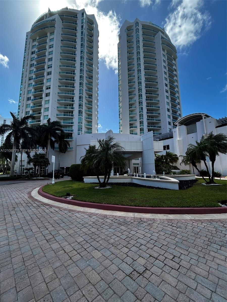 High-rise residential towers featuring glass balconies, a circular paver driveway, manicured landscaping, and a tiered decorative fountain