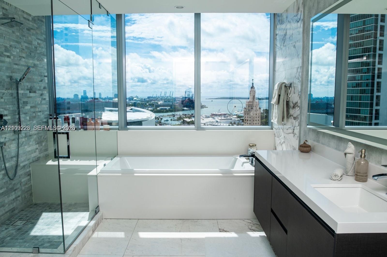 Floor-to-ceiling windows, glass-enclosed walk-in shower with stone tile wall, built-in soaking tub, and dark wood-finish vanity with white countertop