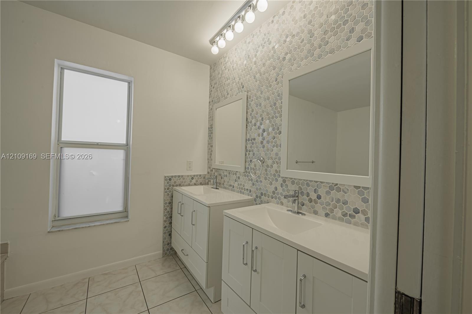 Dual white vanities with integrated rectangular basins, honeycomb-pattern mosaic tile backsplash, framed wall mirrors, and multi-bulb vanity lighting