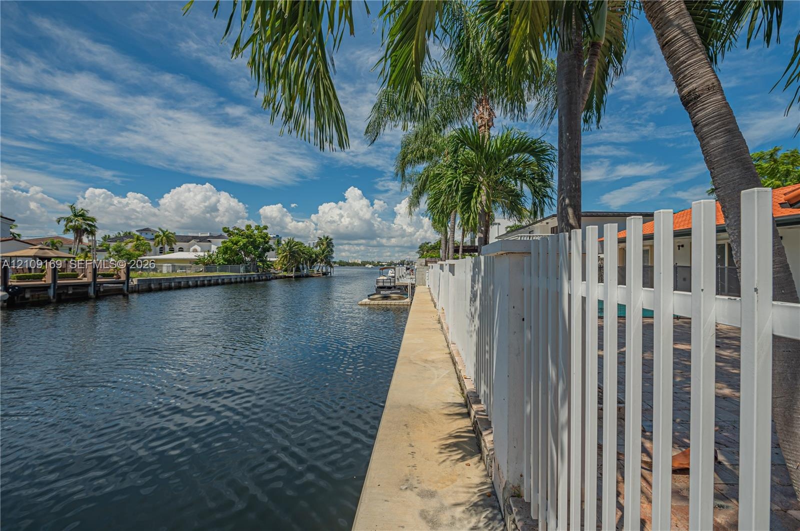 Waterfront property featuring a concrete seawall, white picket fencing, and mature palm trees
