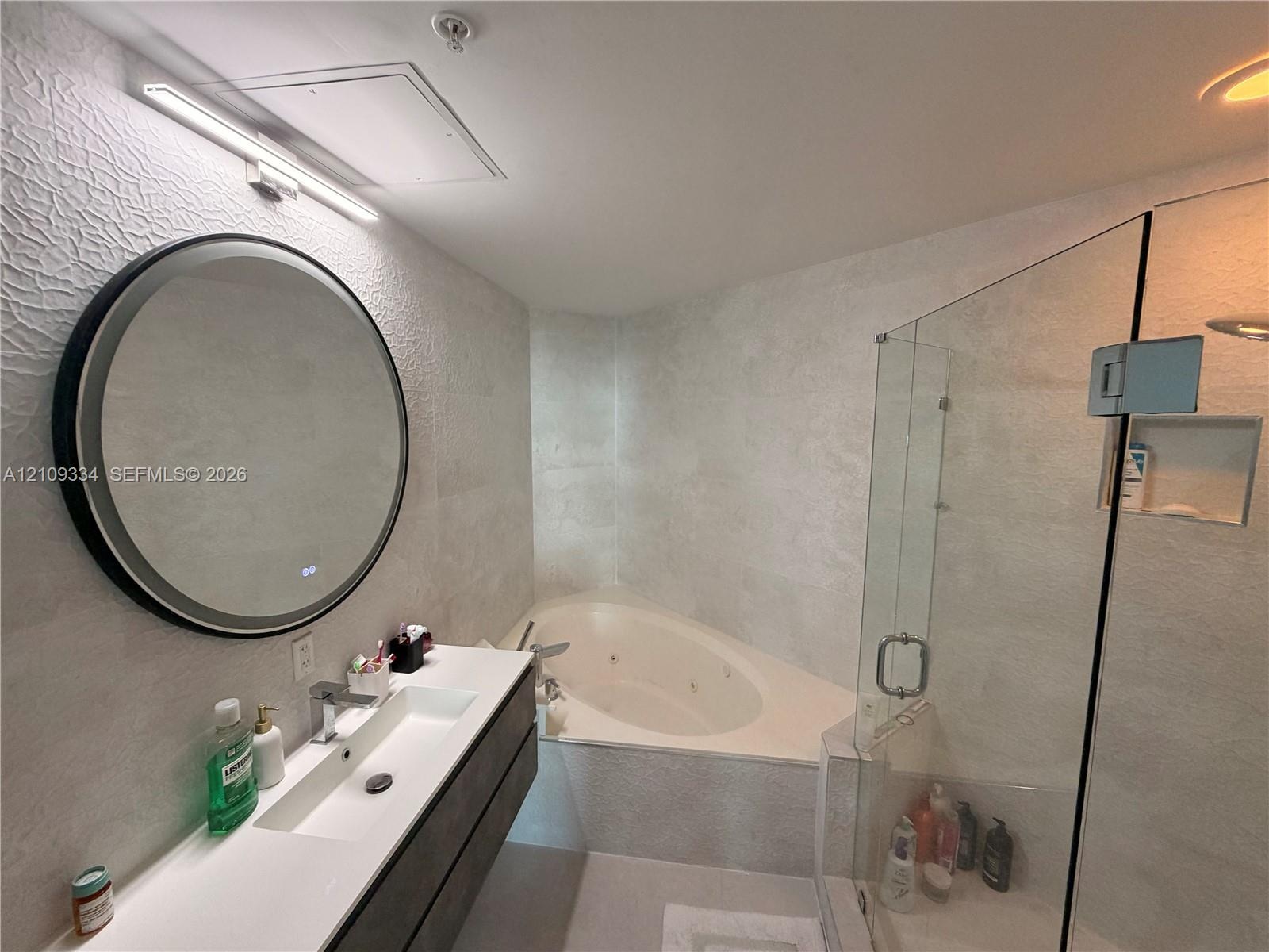Modern floating vanity featuring an integrated basin, circular backlit mirror, glass-enclosed shower, and built-in jetted tub with tiled surround