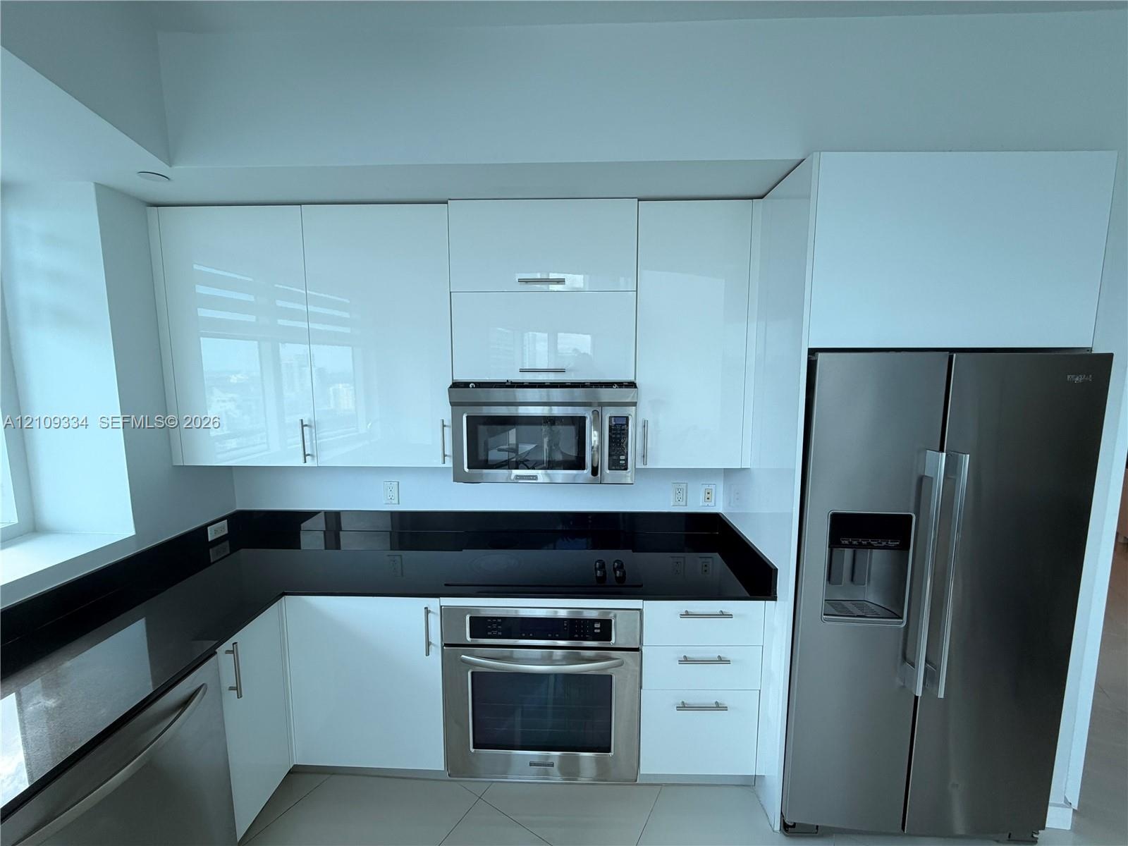 Contemporary kitchen featuring high-gloss white cabinetry, dark stone countertops, stainless steel appliances, and large-format tile flooring