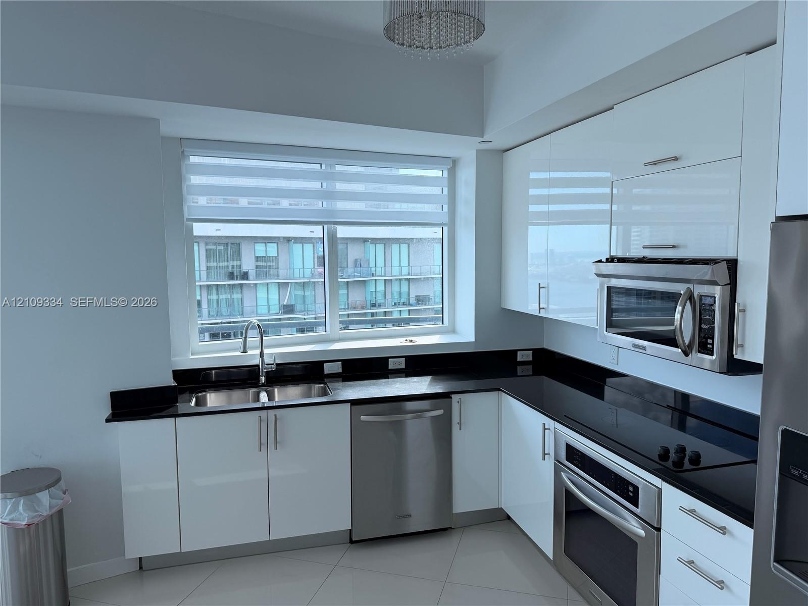 Kitchen featuring high-gloss white cabinetry, dark stone-finish countertops, stainless steel appliances, large double-basin under-mount sink, and a mo