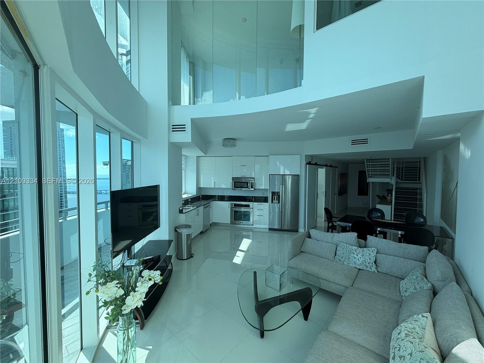 Double-height living area featuring floor-to-ceiling glass windows, high-gloss kitchen cabinetry, stainless steel appliances, and interior glass mezza