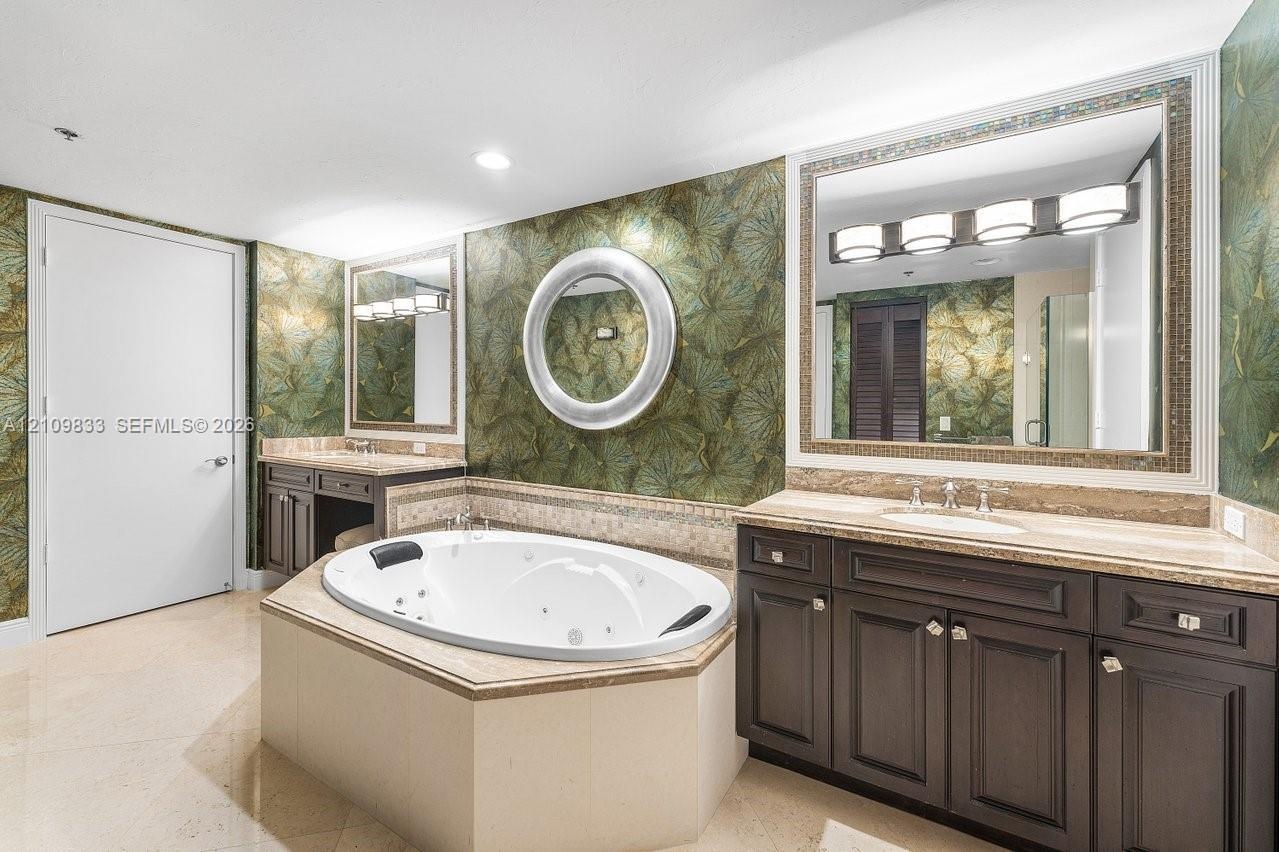 Oval jetted tub with tiled surround, dual dark-finish wood cabinetry, integrated lighting, stone-top vanities with undermount sinks, and wall-mounted 