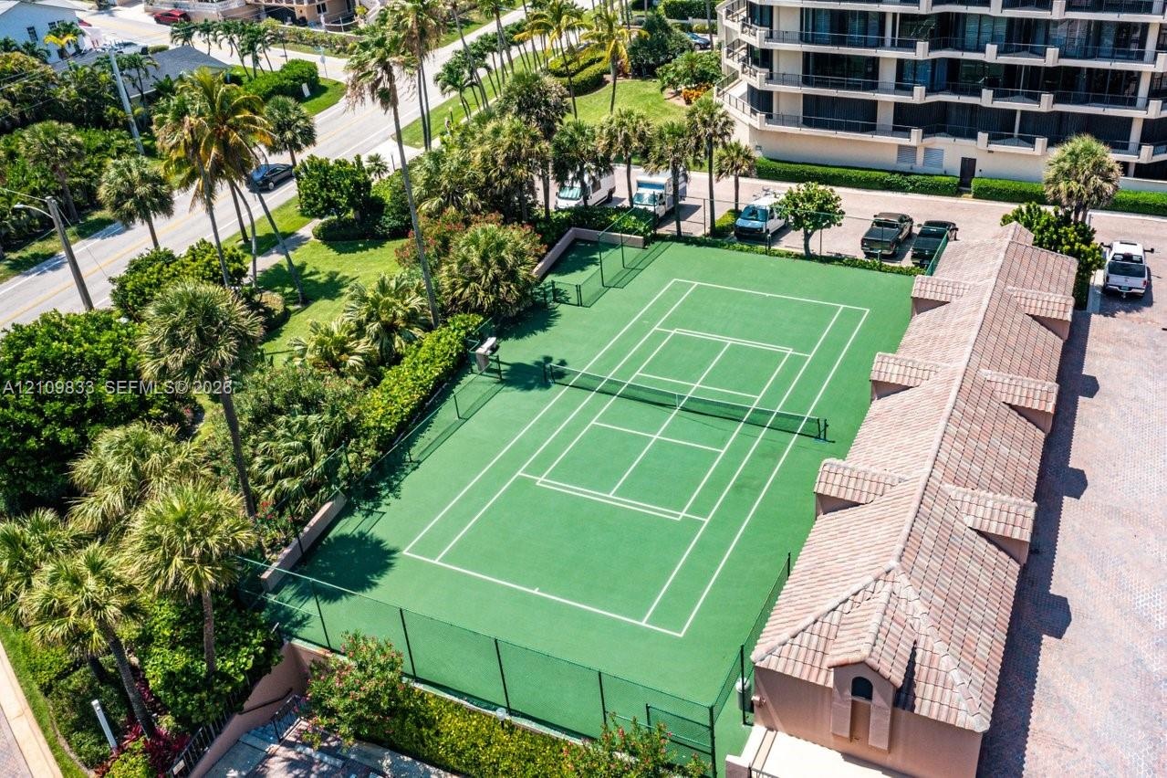 Tennis court featuring a perimeter fence, surrounding mature landscaping, palm trees, and an attached structure with a terracotta-style tile roof